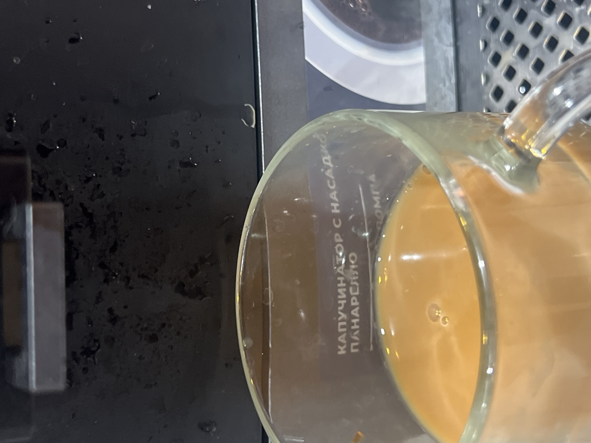 A close-up shot shows a transparent glass mug filled with a light brown liquid, possibly coffee with milk. The mug rests on a black surface, likely part of a coffee machine. Some droplets and stains are visible on the surface. A section of a metal grid pattern is seen in the upper right corner, probably part of the machine's design. The word "КАПУЧИНАТОР С НАСАДКОЙ" is printed vertically on the side of the mug, indicating that the liquid may be a cappuccino. The location is Boguchar, Russia.