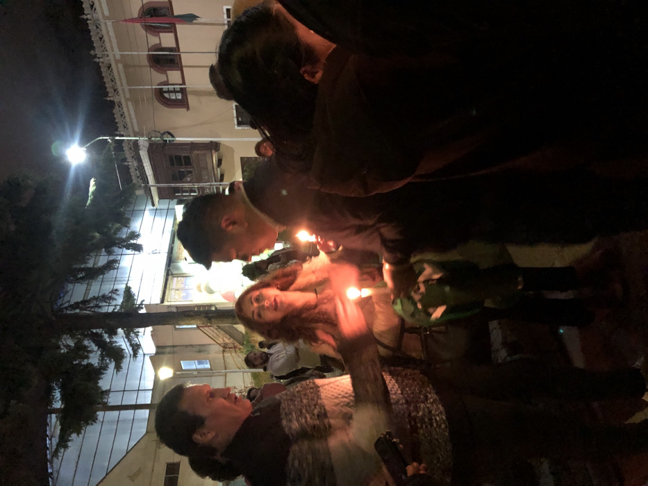 A group of people gather in the dark, illuminated by candlelight. An older woman with short dark hair, wearing a textured sweater, looks up at a younger woman with long red hair, who is holding a lit candle. Behind them, a young man with a short haircut is also holding a candle. The scene is dimly lit, with trees and buildings visible in the background. A street lamp casts a bright light on the scene.