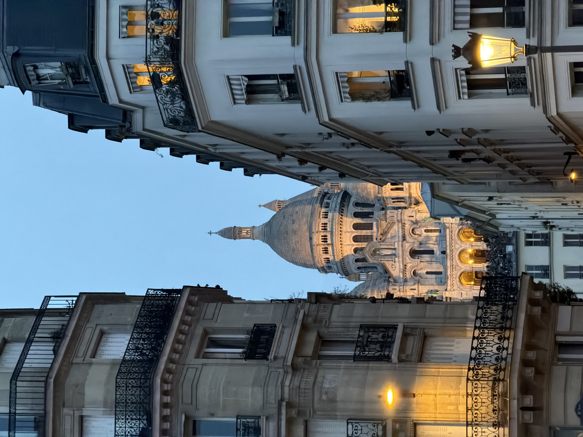 The photograph depicts a view of the Sacré-Cœur Basilica in Paris, France, framed by buildings on either side of the street. The basilica, with its prominent dome, is centrally positioned. The architecture of the surrounding buildings is typical of Paris, featuring windows and ornate balconies. Streetlights illuminate the buildings. The sky is a pale blue.
