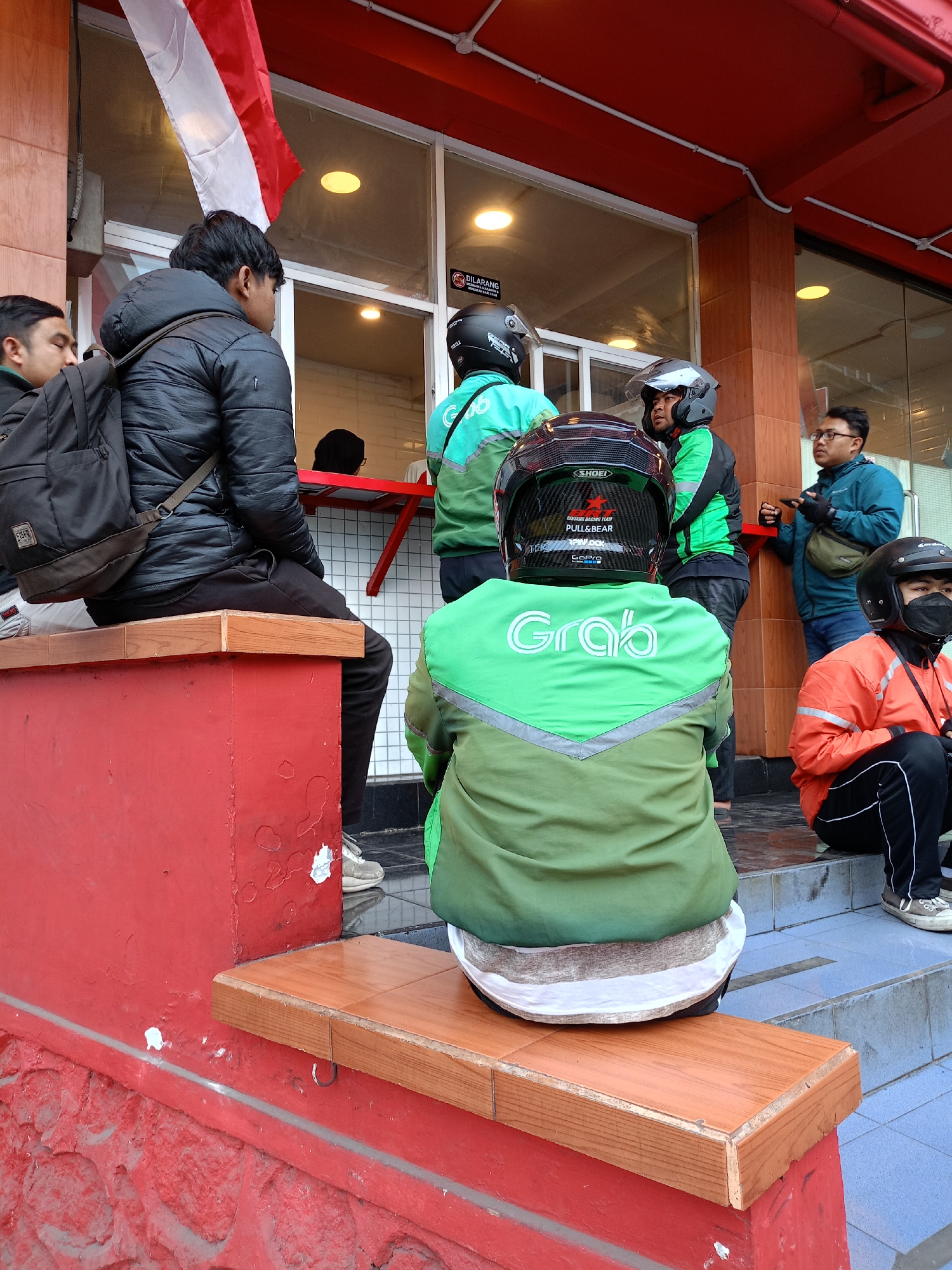The image captures a street scene in Batu, Indonesia, featuring a group of people, likely Grab delivery riders, gathered outside a building with a red facade and a red awning. In the foreground, a person in a green Grab jacket is seated on a wooden bench, facing away from the camera. To their left, another individual in a black puffer jacket and backpack is also seated on a red structure. Several other individuals wearing Grab jackets and helmets are visible standing and sitting on steps leading up to the building entrance. A red and white Indonesian flag hangs prominently above the entrance. The environment suggests a commercial area, possibly a restaurant or food establishment, with tiled floors and glass windows. The overall atmosphere appears casual, with individuals waiting or resting.