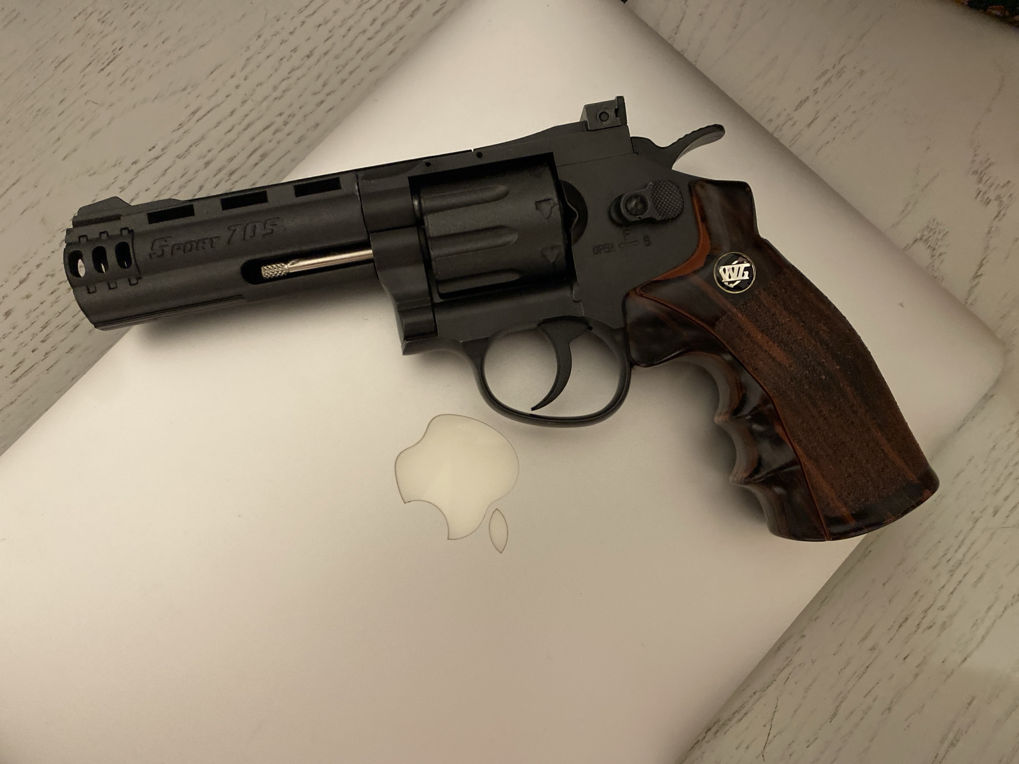 A black revolver handgun with a wooden grip bearing the "WG" logo rests on top of a closed silver Apple laptop. The handgun is angled, with the barrel pointing towards the upper left of the image and the grip towards the lower right. The Apple logo on the laptop is visible beneath the gun. The background is a surface with a light, striated pattern. There is no indication that the image was taken in Sochi, Russia.