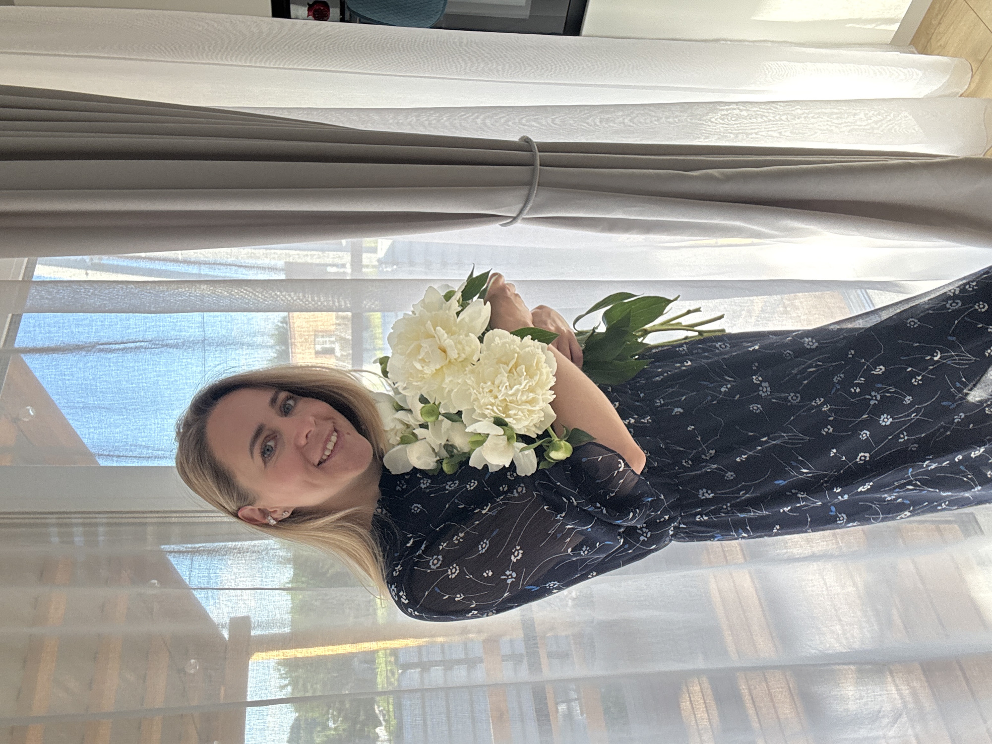A blonde woman wearing a dark blue floral dress smiles as she holds a bouquet of white peonies. She stands indoors by a window with sheer white curtains and gray drapery. Sunlight streams through the window, illuminating the scene and creating a soft glow. The background shows an outdoor view with trees and a building, suggesting a residential area. The woman appears to be in a home environment. The image is taken from a medium shot, focusing on the woman and the flowers.