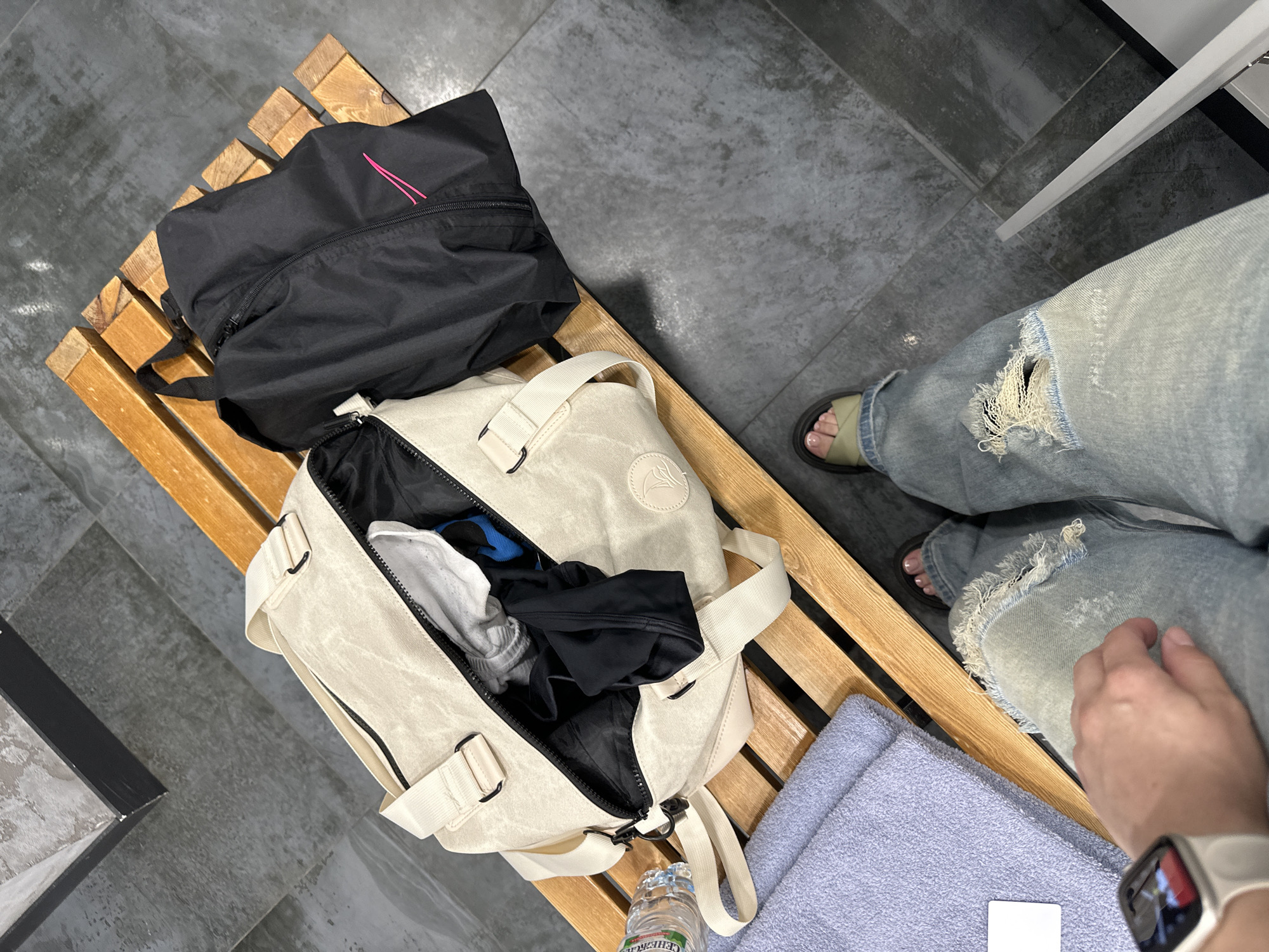 A first-person view shows a wooden bench in what may be a gym or locker room with a black Nike bag on top left, a tan duffel bag with an open zipper on the lower left, a blue towel, a small white card, and a clear plastic bottle of water with a white label. The person taking the picture is wearing light-wash ripped jeans and sandals. The floor is gray tile. The photograph was taken in Perm, Russia, as indicated by the Cyrillic script on the water bottle. The person in the picture is likely preparing to engage in a workout or is packing after a workout. The visible wrist shows a smart watch with a white band.