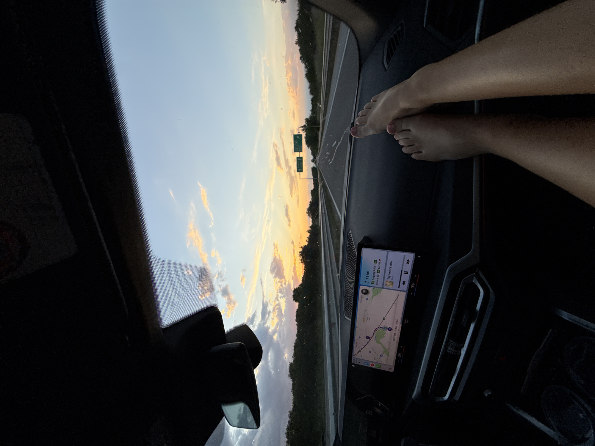 Here is a description of the image:
The image shows a view from inside a car driving on a highway at sunset. Feet are resting on the dashboard. A navigation system displays a map with a route marked to Sofia, Bulgaria from Cherna Gora, Bulgaria. Information indicates the route is 87km long and the ETA is 21:27.