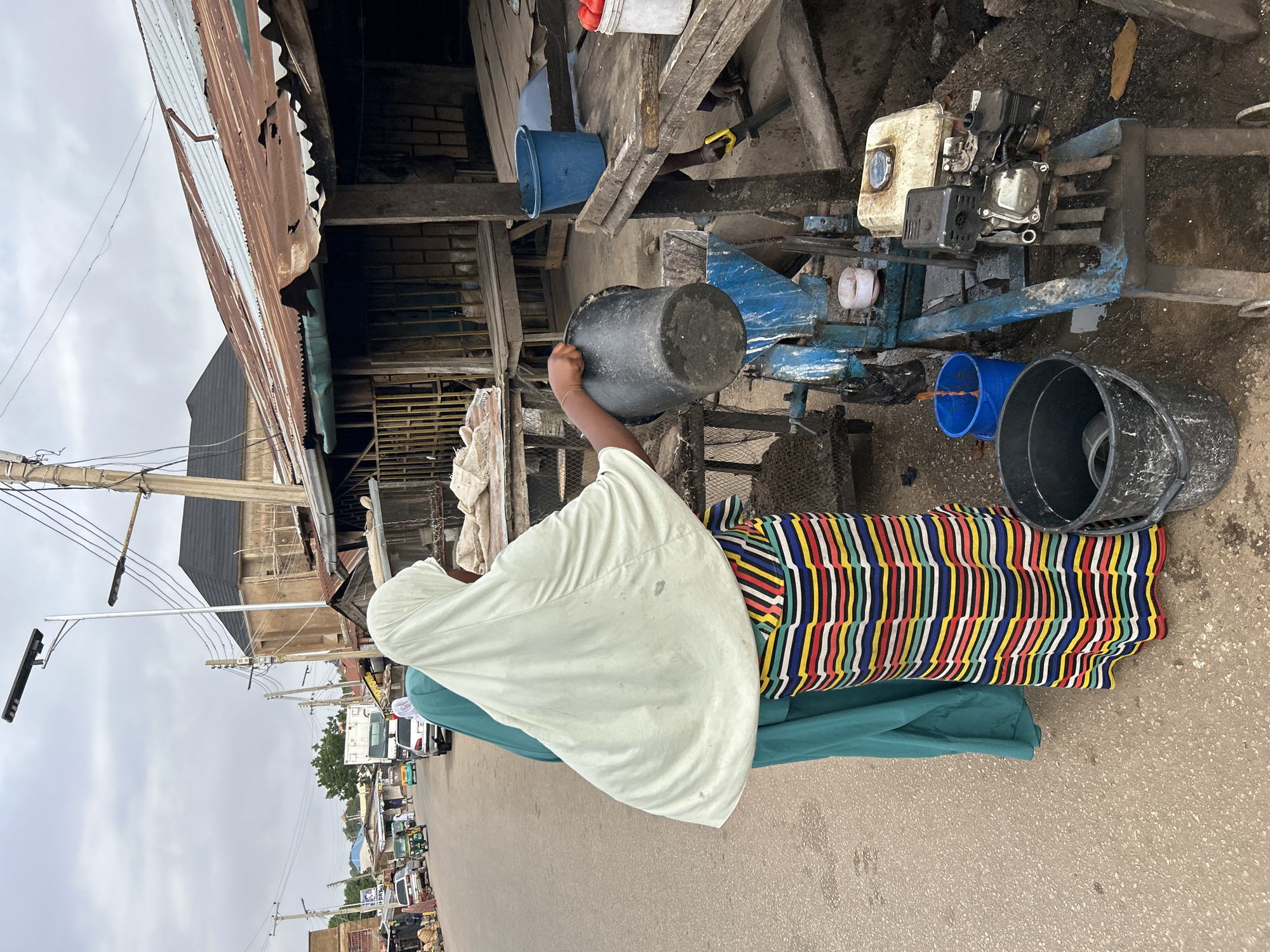 A woman in Jos, Nigeria, wearing a striped, multicolored skirt, a teal underskirt, and a light green head covering, lifts a grey bucket near a mechanical water pump, which is connected to a blue plastic pipe. Two blue and grey buckets rest nearby on the pavement. The woman is situated on a city street near a row of buildings made of corrugated metal and other construction materials. In the background, a busy streetscape is visible with other people, cars, trucks, and tuk-tuks. Electrical poles line the street. The sky is overcast.