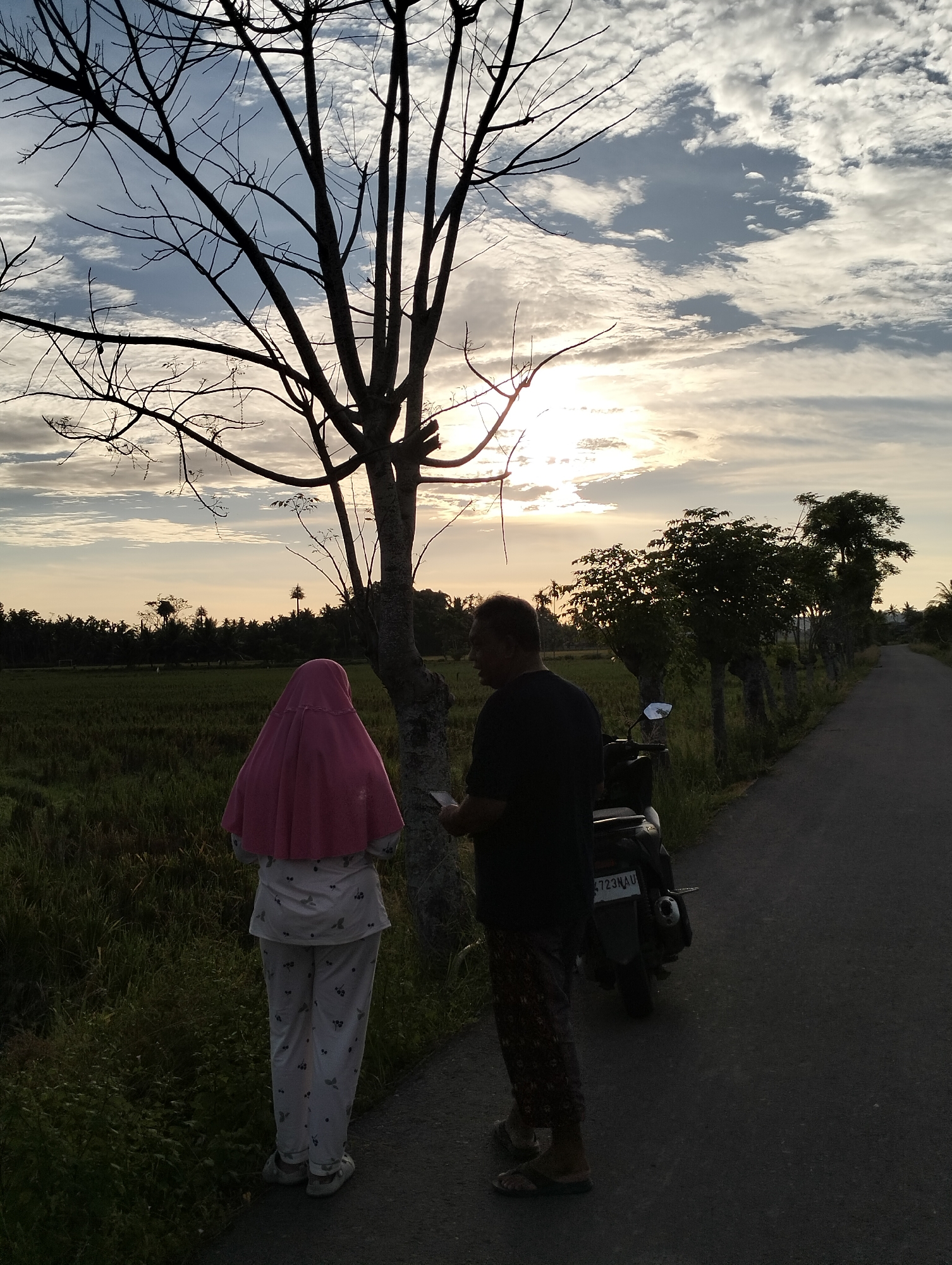 An outdoor scene in Meunasahalue, Indonesia, showing two individuals standing near a tree alongside a road. One person, wearing a pink headscarf and patterned pajamas, stands with their back to the camera. The other, a man, stands facing the first person, holding an object (likely a phone or document). A parked motorcycle is nearby, with a license plate visible (4723KAH). The background includes a rice field, trees, and a cloudy sky with the sun visible. The scene is bathed in soft, natural light, suggesting either sunrise or sunset.