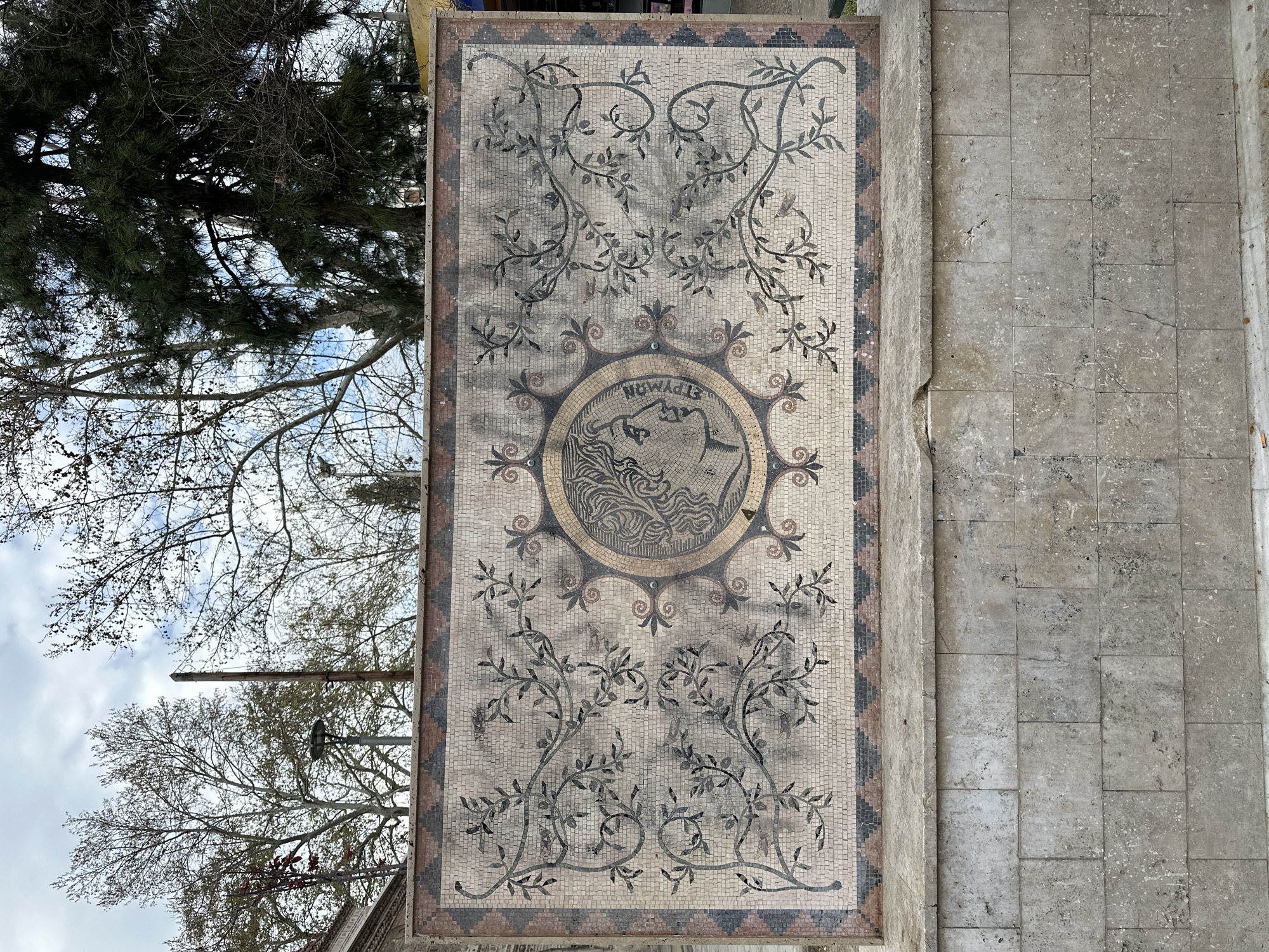 The image shows a mosaic in the city of Serres, Greece. The mosaic is rectangular and features a central medallion with an image of a human figure and the word "ΕΡΥΜΩΝ". The medallion is surrounded by decorative elements including foliage patterns and a border with repeating floral motifs. The mosaic is bordered by a patterned edge of small tiles in varying colors. To the left of the mosaic, out of focus, are trees and sky. To the right are gray stone tiles.