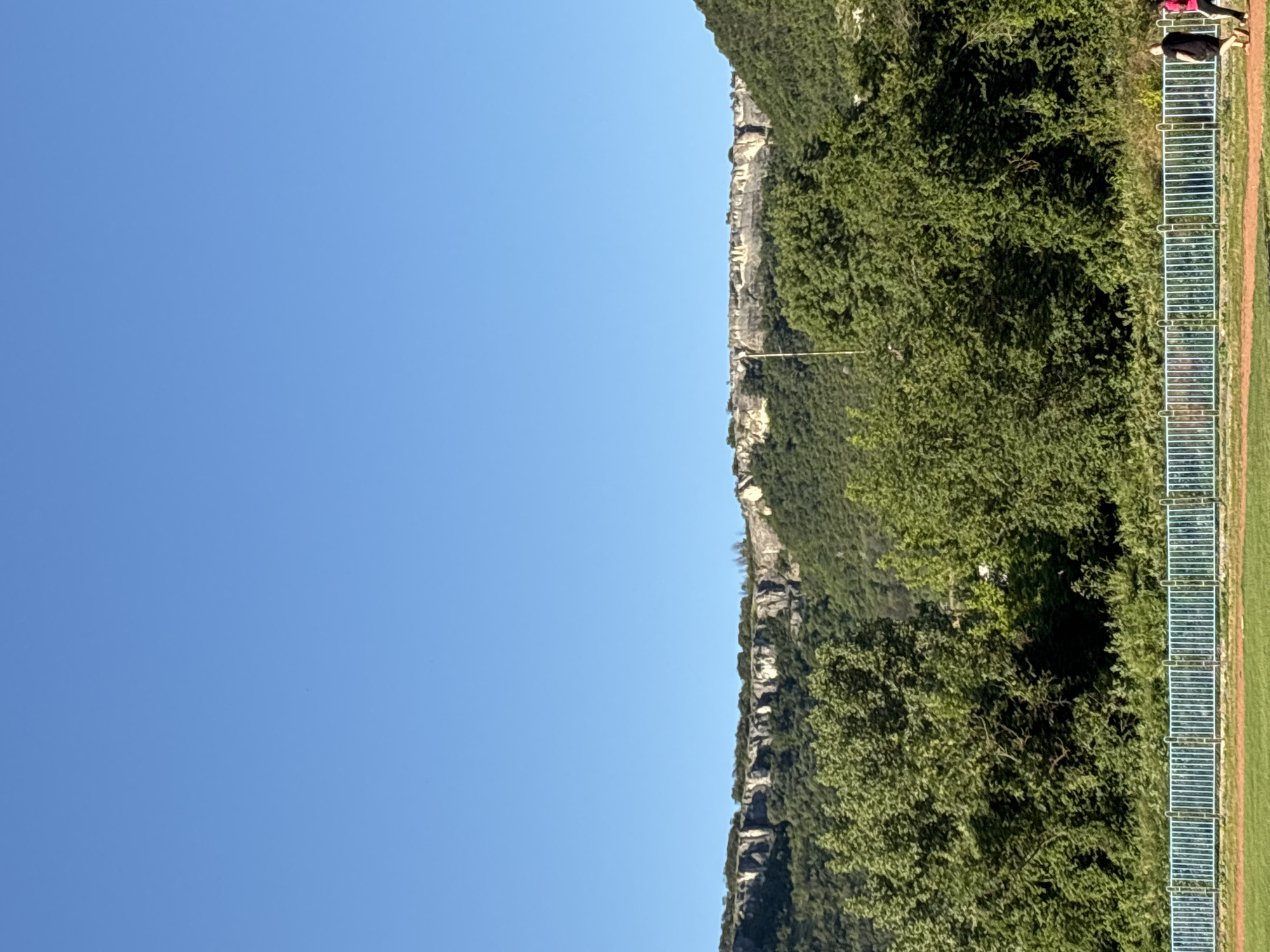 This is a vertical, outdoor, daytime photograph taken in Provadia, Bulgaria. The image features a clear blue sky dominating the upper portion, with a wide shot of a mountain range in the mid-ground. The mountains are covered in lush green trees, with a prominent, rugged cliff face visible along the ridge. In the foreground, there is a blue metal fence bordering a red track or field. Two individuals are walking away from the camera along the track, on the right side of the frame. One person is wearing a black t-shirt and shorts, and the other is wearing a pink t-shirt and dark shorts. The overall scene depicts a natural landscape with a man-made recreational area.
