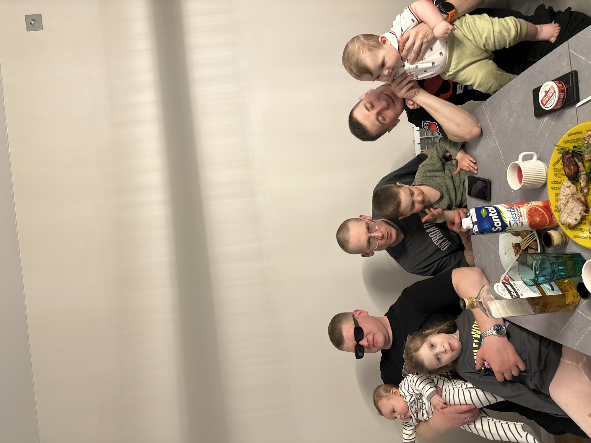 An indoor scene captures a multi-generational group of seven people gathered around a gray table, likely for a meal or social occasion in Chaikovsky, Russia. The image appears to be taken from above, with the subjects looking up or around.
On the left side of the frame, an adult man with dark hair holds a baby with light hair, both looking towards the camera. Next to them, a young boy with light hair is seated, looking down at the table. On the opposite side, an adult man with a bald head and glasses is visible, looking towards the left. In the lower part of the image, another adult man wearing sunglasses holds two children: a young girl with light brown hair, wearing a black top, who looks directly at the camera, and another baby in a striped outfit, also looking towards the camera. The atmosphere appears relaxed and familial.
The table is laden with various items. A yellow plate with text "PIZZA," "POMODORO," and "paperont" features grilled meat and herbs. Other items include a white mug with red liquid, a carton of "Santal Siesta КРАСНЫЙ Сицилийский апельсин" (Red Sicilian Orange) juice, a bottle of "BUSHMILLS THE ORIGINAL" Irish whiskey, and a blue glass. A metal can with "SIBERIA" and "OFFROAD" text, likely related to snuff, is also visible. Several smartphones are scattered across the table. The background consists of a plain, light-colored wall with a visible electrical outlet.