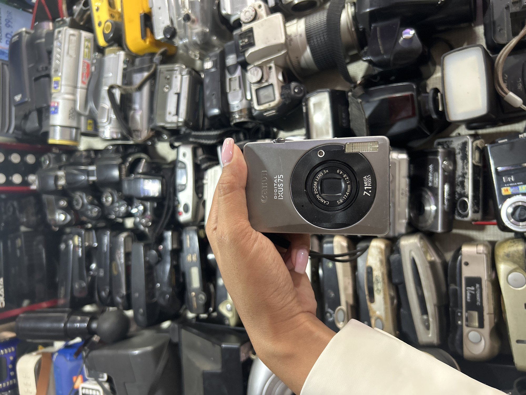 The image shows a close-up of a person's hand holding a Canon digital camera, identified as an IXUS 75 model. The camera has "7.1 MEGA PIXELS" and "CANON ZOOM LENS 3x" printed on its lens barrel. The background is a cluttered display of various electronic devices, predominantly older model cameras and camcorders, suggesting a retail or storage environment. Among the visible items are several compact point-and-shoot cameras stacked vertically, and larger video cameras. The lighting appears to be artificial, casting shadows and highlighting the textures of the devices. There are no explicit geographical markers or people other than the hand holding the camera. The image is cropped to focus on the camera and the hand, with the surrounding items serving as a backdrop. The context of Denpasar, Indonesia is not directly evident from the visual information within the image itself.