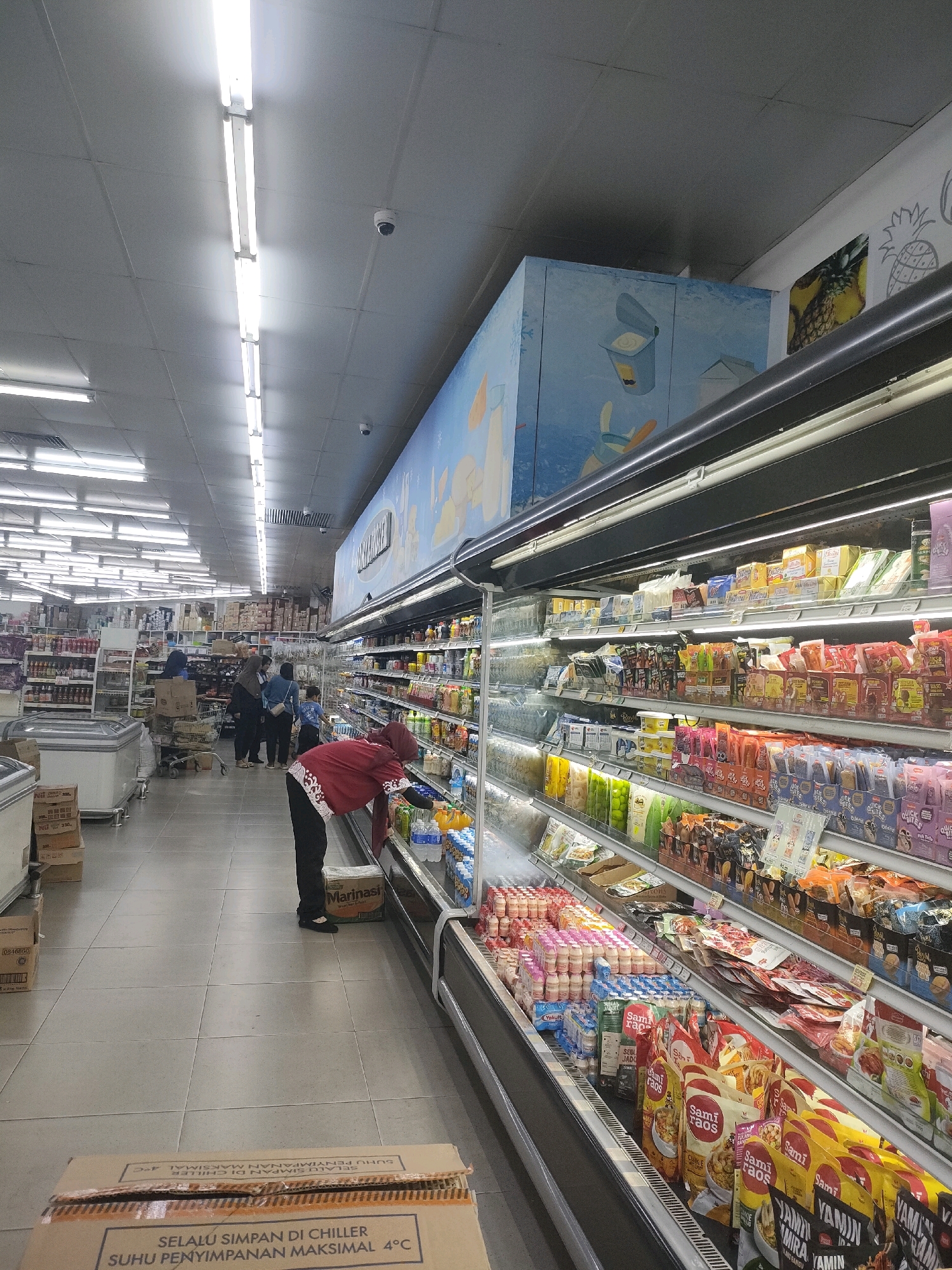 This is a photograph taken inside a brightly lit supermarket in Batam, Indonesia. The perspective is from behind a person wearing a red hijab and a red patterned top, who is reaching into a refrigerated display case. The display case is filled with various food items, including bottles of drinks, yogurt containers, and packaged snacks. To the left of the person, there are more refrigerated units and shelves stocked with products. In the background, several other shoppers are visible, some looking at shelves and others pushing shopping carts. The ceiling is lined with fluorescent lights, and there are security cameras mounted on the ceiling. The floor is tiled, and there are cardboard boxes on the floor in the foreground. The overall impression is of a busy retail environment.