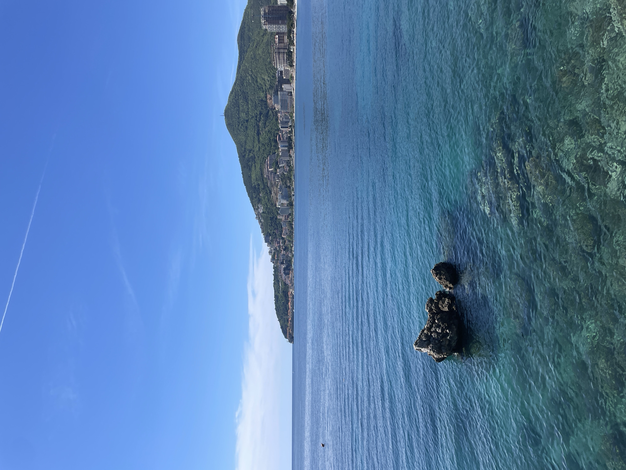 The image shows a coastal view in Budva, Montenegro. The scene features the Adriatic Sea, which is a vibrant turquoise and blue. Two rock formations are visible in the sea, close to the camera perspective. Along the coastline, there is a mountain covered in lush green vegetation, extending into the clear blue sky. Buildings and structures are visible along the base of the mountain where it meets the coast. A faint contrail cuts across the top left of the blue sky.