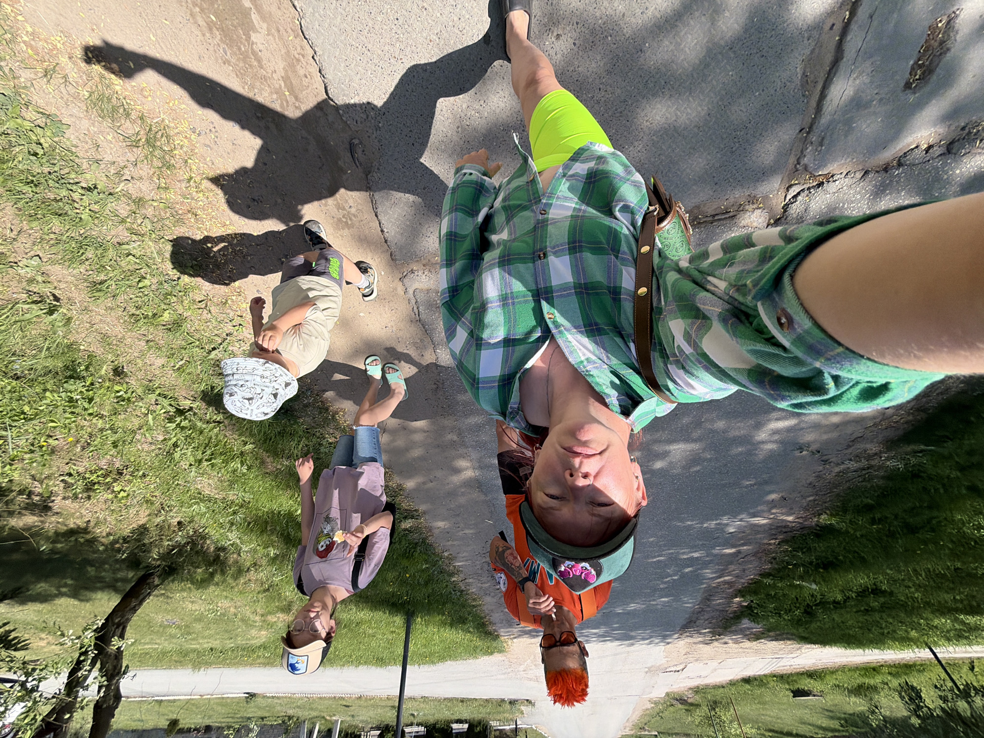 The image is a selfie taken at an elevated angle, showing four people on a paved path next to a grassy area. In the foreground, a person wearing a green and white plaid shirt and bright yellow shorts stands on the path. To the left, two children are standing on the grass. One child is wearing a tan shirt, grey shorts, and a patterned hat. The other child is wearing a lilac t-shirt, jeans, and a baseball cap. In the background is a person wearing an orange shirt and a baseball cap with orange hair. The location is Chaikovsky, Russia.