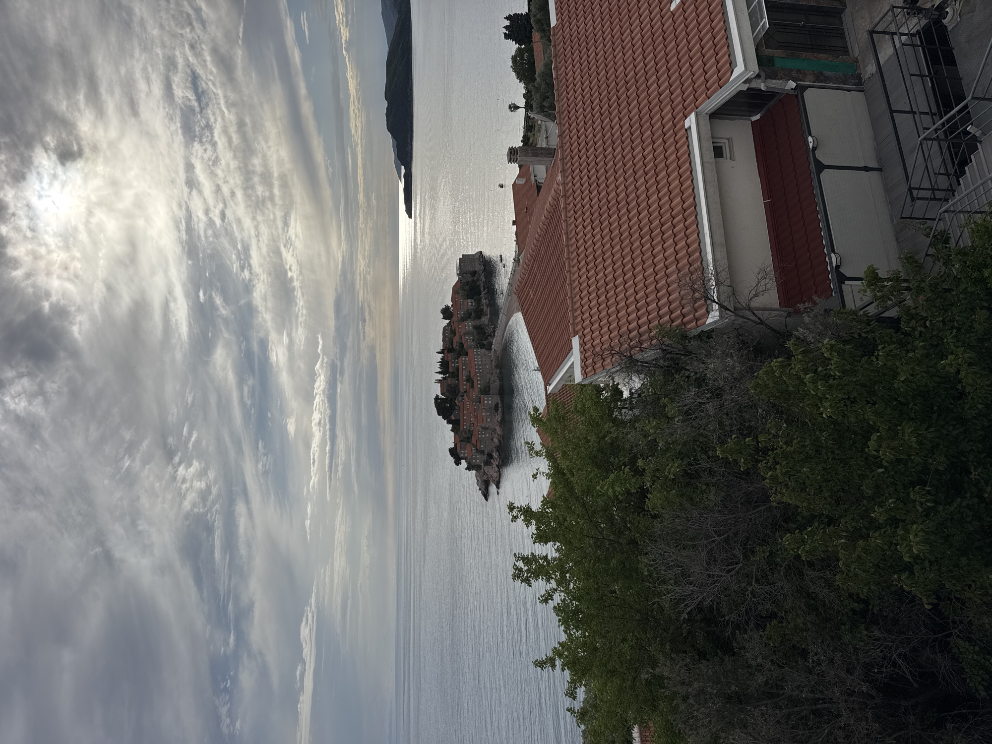 The image captures a view of Sveti Stefan, Montenegro, from an elevated position, possibly a building. The iconic island-town of Sveti Stefan is centrally positioned in the frame, emerging from the Adriatic Sea. The water reflects the sunlight, creating a shimmering effect. The sky is filled with striated clouds, suggesting a dynamic weather pattern.
To the right of the frame, the red-tiled roof of a building is visible. Below the roof, there is lush green foliage from trees and bushes. To the left, the open sea stretches towards the horizon, with a dark landmass visible in the distance.