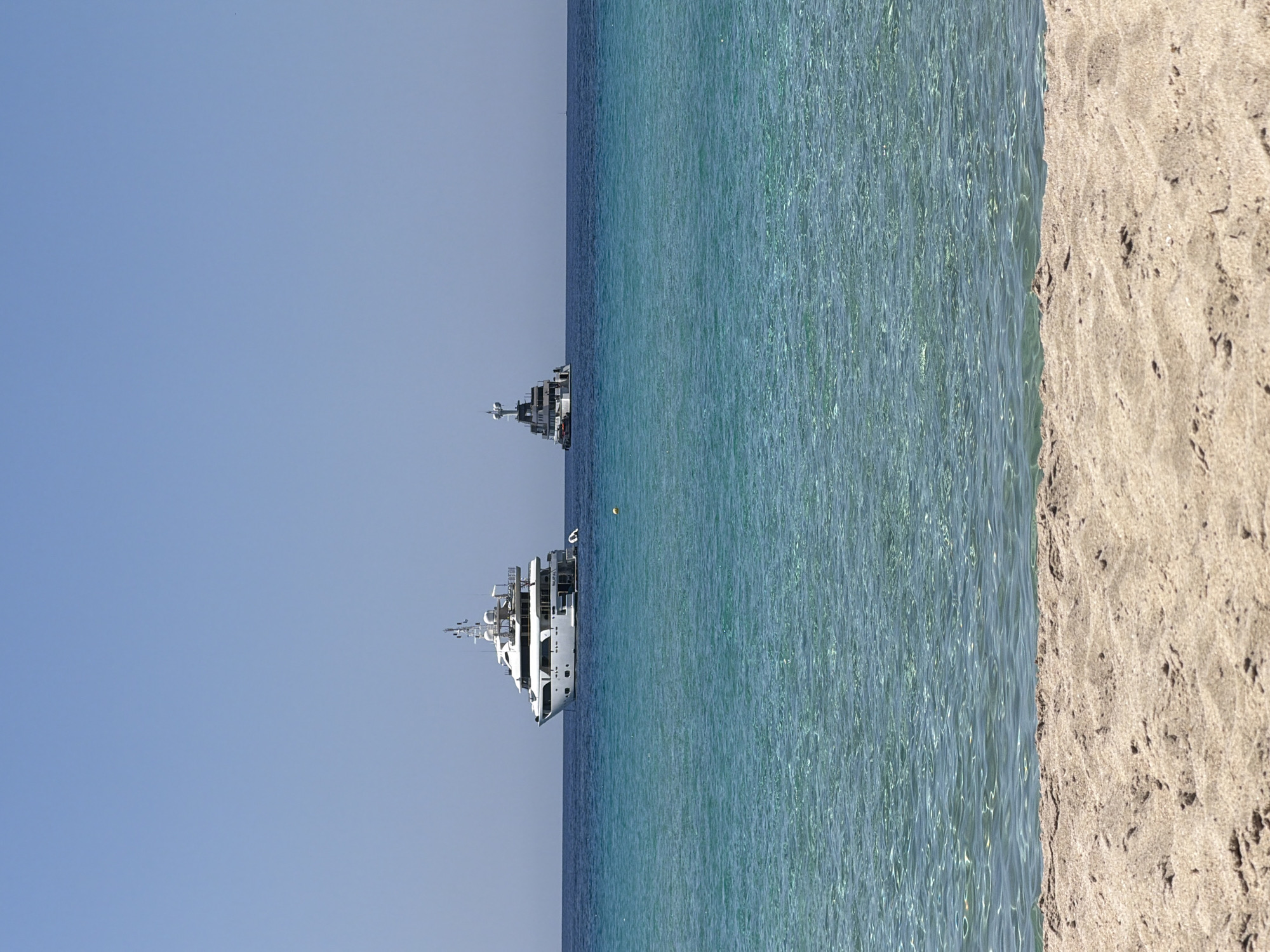 The image shows a clear blue sky over a tranquil turquoise sea. Two large white yachts are anchored in the water, one significantly larger than the other. The larger yacht, on the left, is multi-decked with numerous windows and antennas. The smaller yacht, on the right, is also multi-decked and appears to be a chase boat or support vessel. In the foreground, a sandy beach with textured sand is visible, hinting at a coastal location. The water is calm and clear, allowing visibility of the seabed in some areas. The scene suggests a peaceful day at sea, likely in a Mediterranean setting given the clear water and sunny weather. The location is described as Unknown, Greece, but the visual elements are consistent with many Greek island coastlines. There are no people visible in the image.