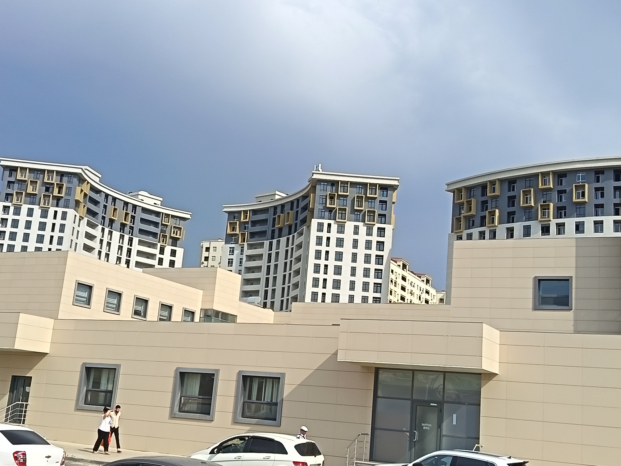 This is a horizontal, medium-angle, outdoor photograph taken in Masazir, Azerbaijan, featuring a modern architectural complex. The sky above is a hazy light blue with some clouds. In the foreground, a low-rise, light-colored building with multiple windows runs across the frame. Two individuals are walking on a sidewalk in front of this building. The male is wearing a light-colored shirt and dark pants, carrying a red bag. The female is wearing a white top and dark pants. To the left, the back of a white car is visible.

Behind the low-rise building, several multi-story apartment buildings are visible. These buildings exhibit curved facades and a distinctive pattern of light and dark grey panels with protruding rectangular window frames, some in a yellow accent color. The architectural style is contemporary, with varying heights and forms. The overall impression is one of a newly developed urban or suburban area.