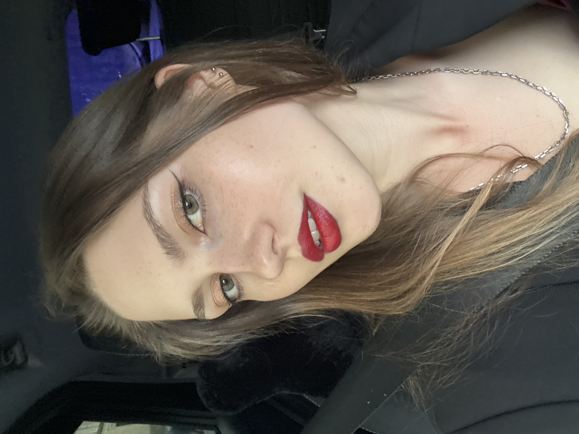 This is a close-up, vertically oriented, portrait-style photograph of a young woman. She has light brown hair, fair skin with freckles, and light-colored eyes. Her makeup includes winged eyeliner and bright red lipstick. She is wearing a silver chain necklace, and a small stud earring is visible in her left ear. A faint red mark, possibly a hickey, is visible on her collarbone. The background appears to be the interior of a vehicle, with dark upholstery and a hint of a blue object behind her. The lighting suggests it is daytime, but the exact time and weather conditions are not discernible. The woman's expression is neutral or slightly suggestive, with her head tilted. There is no visible text in the image. The overall impression is that of a selfie taken in a car.