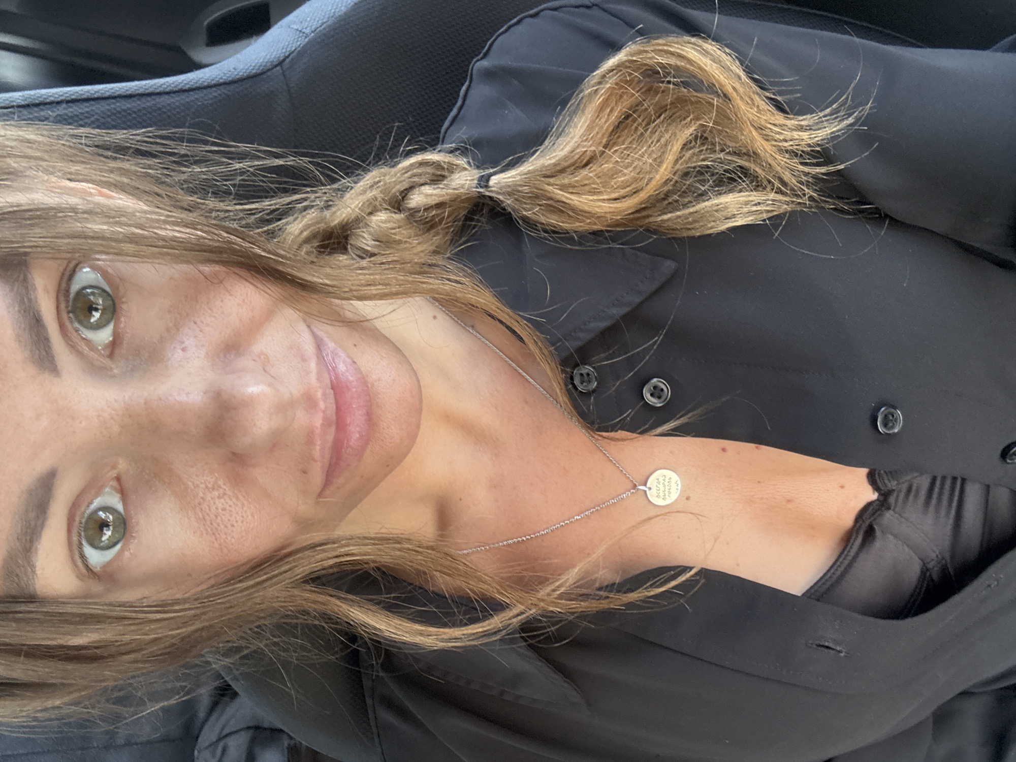 A woman with light brown hair, partly braided, and hazel eyes is positioned in what appears to be a car seat. She is wearing a black shirt with a black undergarment visible at the collar. She also wears a silver necklace with a round pendant. The photograph is taken from an upward angle. No other individuals or objects are clearly visible. The location is not discernible from the image.