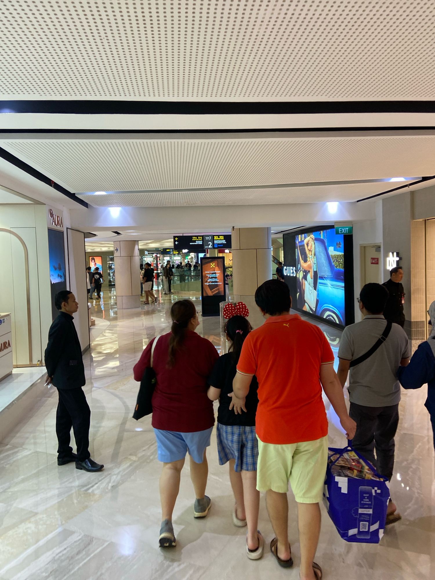 In a well-lit, modern indoor walkway, likely within a shopping mall or airport in Surabaya, Indonesia, a group of people are walking away from the camera. The walkway features a perforated white ceiling and marble-like flooring that reflects the overhead lights.
On the left, a man in a black suit is walking towards the camera, looking to his left. Further down the walkway, a family group is walking away. The woman in the foreground is wearing a maroon top and blue shorts. She is followed by a child in a black shirt and blue plaid shorts, and then an adult male in an orange t-shirt and light green shorts. The man is carrying a large blue shopping bag filled with various items. To the right of this group, another man in a grey t-shirt and dark pants walks with a black bag slung over his shoulder.
In the background, several digital screens display advertisements and directional information. One large screen shows a fashion advertisement for "GUESS" featuring a woman and a car. An "EXIT" sign is visible above a doorway on the right. Other individuals can be seen further in the distance, suggesting a busy public space. The overall atmosphere is one of transit and commercial activity.