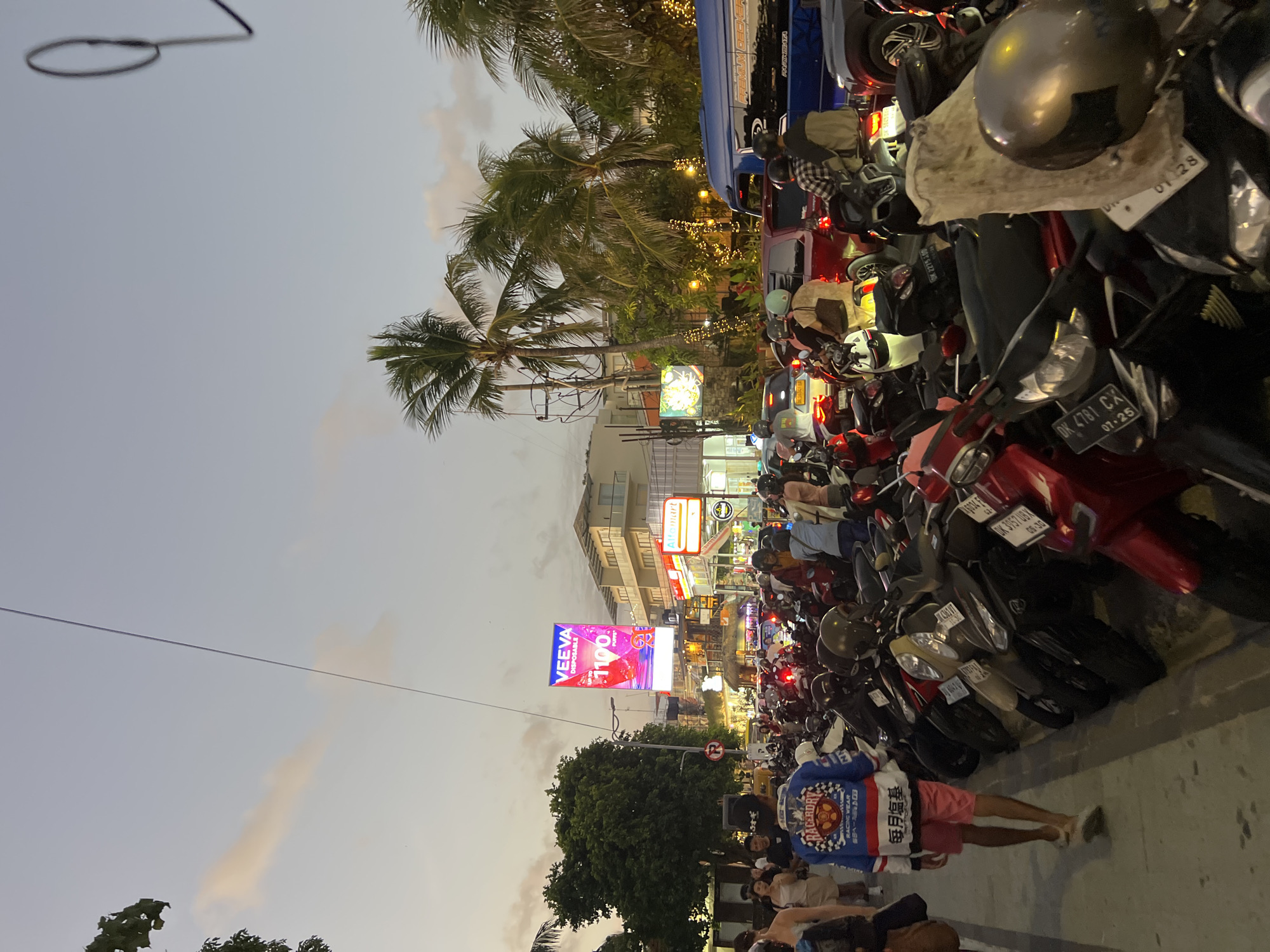 The image depicts a busy street scene in Kuta, Indonesia, likely during the evening or dusk. A dense crowd of people and a large number of parked motorcycles fill the foreground and middle ground. The atmosphere suggests a lively commercial or tourist area.
In the foreground, a young man wearing a blue and white racing jacket and pink shorts is walking away from the camera. He is surrounded by numerous parked motorcycles, indicating a popular parking area. The motorcycles are of various colors and models, and many have license plates visible.
Further back, the street is congested with more motorcycles and some cars. People are seen walking and riding their vehicles, contributing to the sense of activity. Buildings line the street, some of which appear to be shops or restaurants, with illuminated signs. One prominent sign displays "VEEVA" and the number "1100". Another sign reads "Alfamart". Palm trees are visible in the background, characteristic of a tropical location. String lights are also present, adding to the ambiance.
The sky is a pale blue with hints of orange and pink, typical of a sunset or twilight. A power line stretches across the upper portion of the image. The overall scene is one of vibrant street life, with a strong emphasis on motorcycle culture and commercial activity in Kuta, Indonesia.