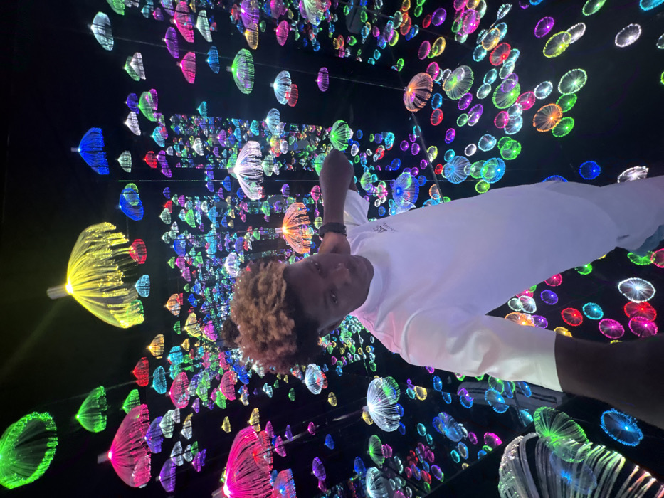 A young male is depicted standing within a dark, highly reflective art installation in Jakande, Nigeria. The individual, possessing medium-dark skin and short, curly hair with a lighter blonde/brown top section, is centered in the frame. He wears a plain white long-sleeved shirt with a small dark logo on the left chest and matching white trousers. A small nose stud is visible on his left nostril, and a dark-colored watch or bracelet is present on his left wrist. His right arm is bent with his hand placed behind his head. He maintains direct eye contact with the camera. The surrounding environment is characterized by hundreds of suspended, illuminated fiber-optic light fixtures, shaped like small umbrellas or jellyfish, emitting a wide spectrum of colors including green, yellow, pink, blue, red, purple, turquoise, and orange. These lights are distributed at various depths and heights against a dark background, creating an impression of infinite space due to highly reflective surfaces on the floor and potentially the walls.