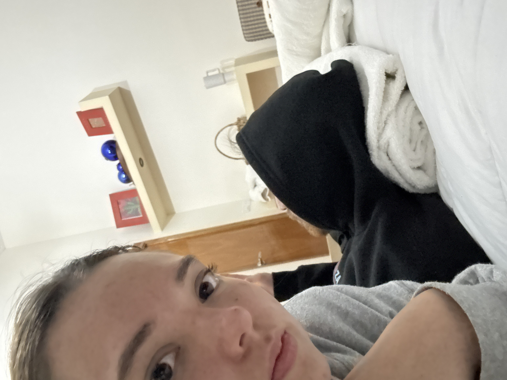 A close-up selfie shows a fair-skinned person with brown eyes and light brown hair, lying down and looking directly at the camera. Behind this person, another individual is visible wearing a black hooded sweatshirt. Piled next to the hooded figure are rolled-up white towels and a larger white item that could be a sheet or blanket.
In the background, a light-colored wall is visible, along with a wall-mounted shelf or decorative item featuring blue spheres and framed pictures. A towel rack and a small piece of wooden furniture are also discernible.
The location is not discernible from the image to pinpoint a specific area in Puebla, Mexico.