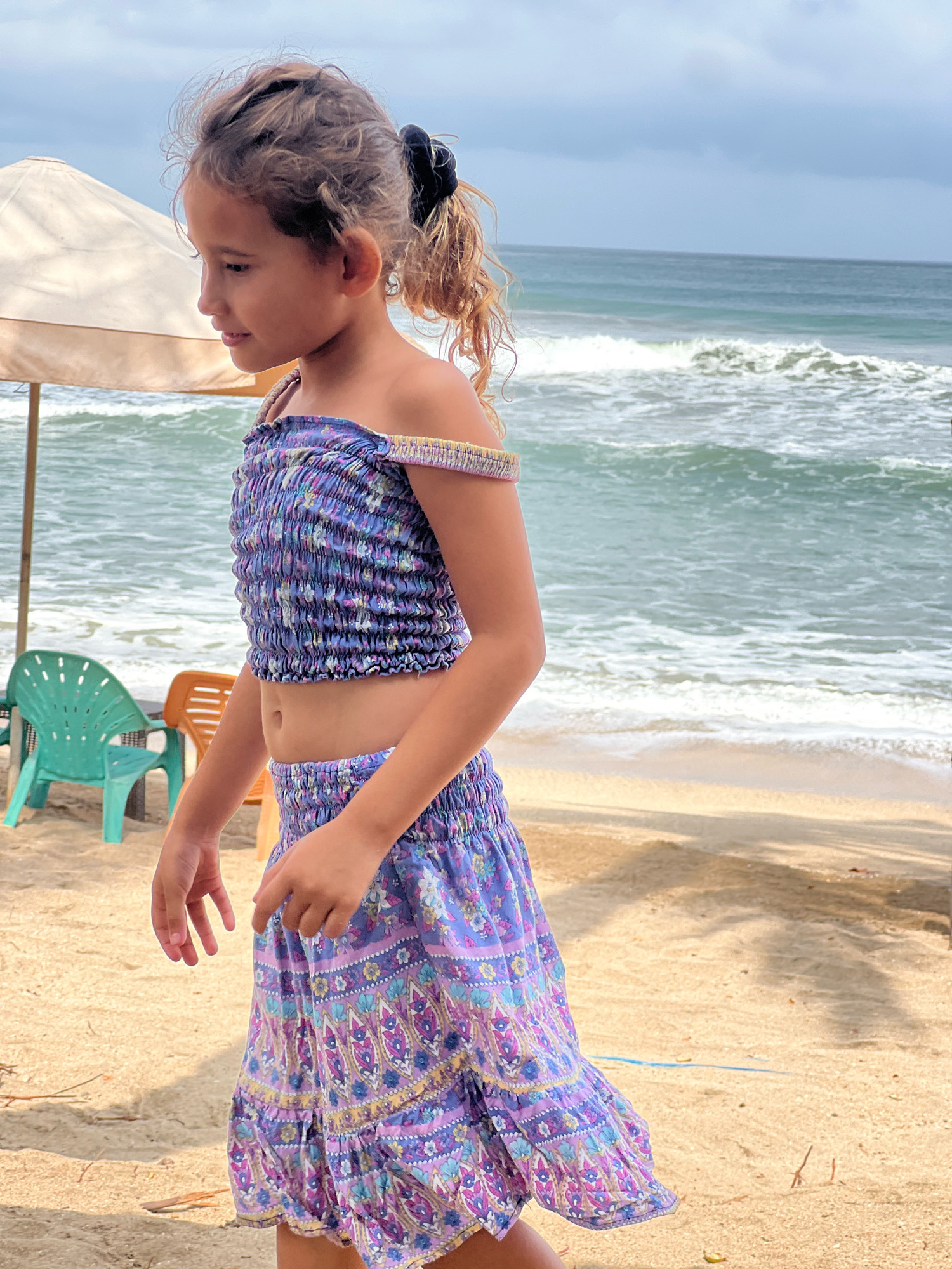 A young girl with blonde curly hair tied back in a ponytail stands on a sandy beach in Kuta, Indonesia. She is wearing a purple and blue floral-patterned off-the-shoulder crop top and a matching skirt. The ocean with moderate waves is visible in the background, with a cloudy sky above. To the left of the girl, there is a beige beach umbrella and a green plastic chair. The overall scene depicts a casual day at the beach.