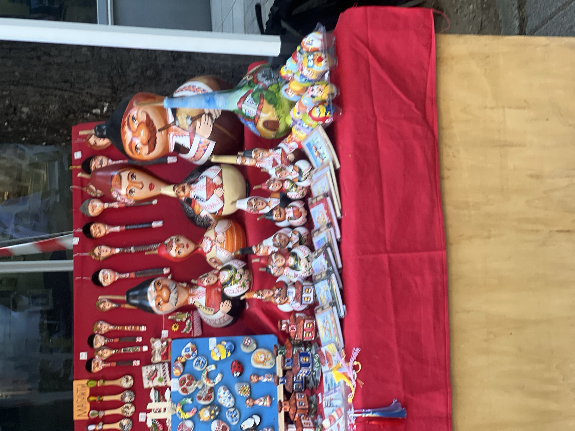 A close-up, eye-level shot shows a market stall display in Plovdiv, Bulgaria. Wooden spoons and other small, hand-painted items are arranged on a red cloth. The spoons feature human faces, traditional Bulgarian costumes, and miniature scenes. Additionally, there are small painted stones and miniature houses. The background includes a building exterior.