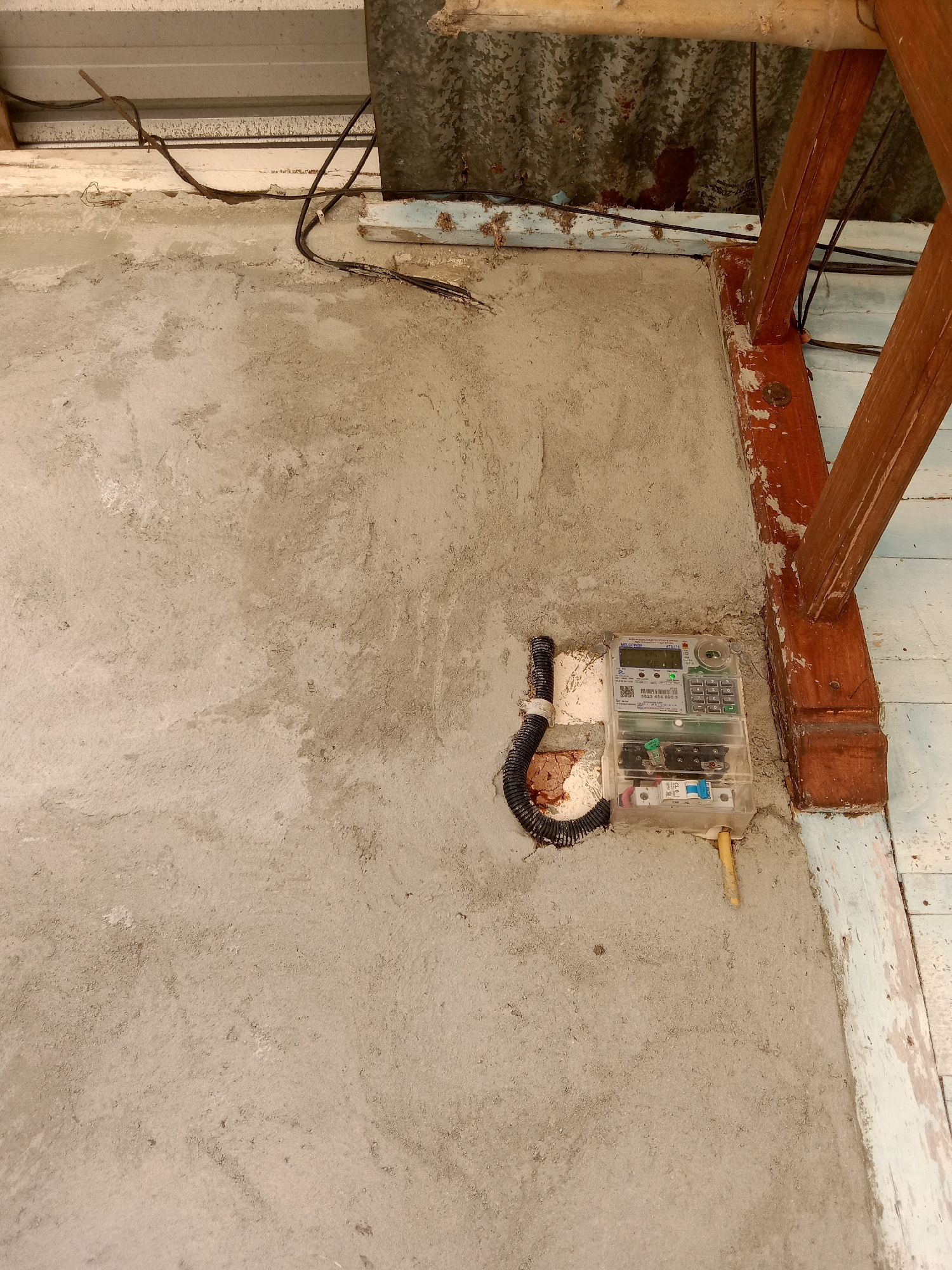 An outdoor view of a concrete wall with an electricity meter attached. The meter is housed in a clear plastic casing and displays a digital readout. A black, corrugated conduit runs from the wall to the meter. To the right of the meter, a wooden support beam is visible, part of a structure that appears to be outdoors. The background shows a corrugated metal roof and some wooden planks. The scene is located in Stiaji, Indonesia.