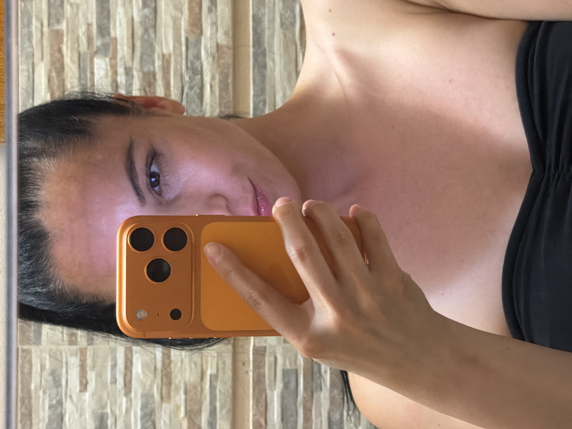 A woman is taking a selfie in a mirror. She is holding an orange smartphone in her right hand. The background appears to be a tiled wall. Her left shoulder and part of her chest are visible. The image does not provide any information about the location in the city of Pleven, Bulgaria.