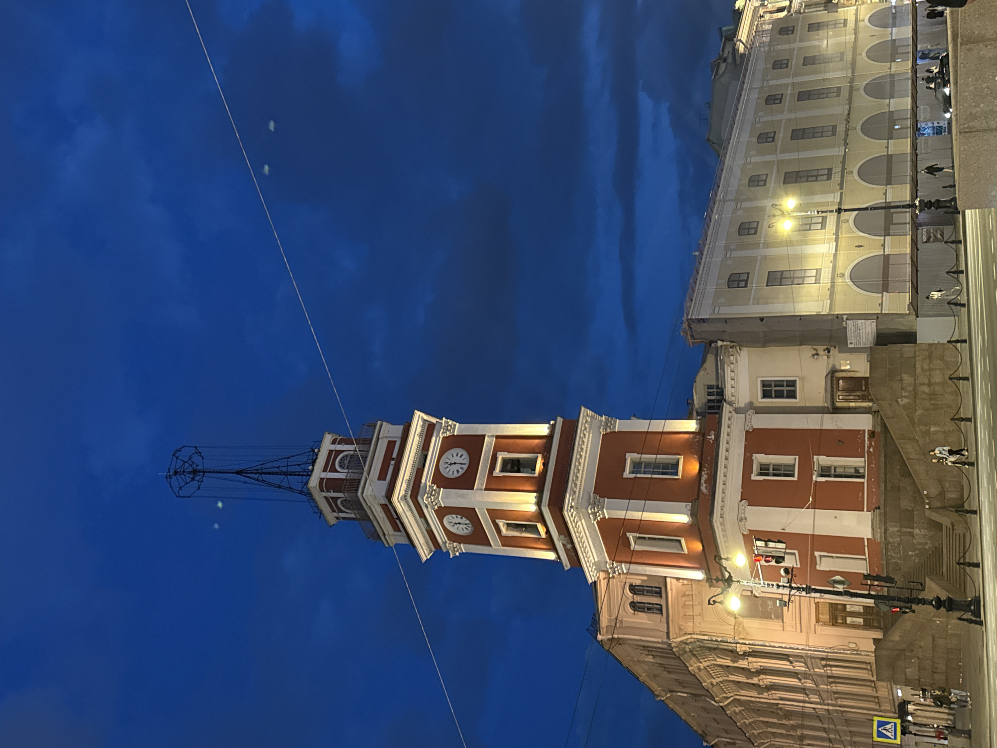 The photograph, captured in Saint Petersburg, Russia, depicts an evening cityscape with the prominent City Duma Tower on the left, illuminated against a dark blue sky. The tower features multiple tiers, painted in a distinctive red and white, adorned with decorative architectural elements and several clock faces. Its apex is crowned with an intricate black metal spire or weather vane structure. The deep blue sky above suggests late evening or night, with faint cloud formations. In the upper left portion of the sky, two small, hazy, bright spots are visible, consistent with lens flares or reflections rather than physical objects.
Adjacent to the tower and extending to the right is a large, multi-story building with a light-colored, possibly cream or yellow, facade. This building is largely obscured by a transparent white netting or scaffolding, indicating ongoing construction or renovation. Warm light emanates from its windows and from streetlights positioned along the street, illuminating the architecture. The building exhibits arched windows on its lower visible levels and rectangular windows higher up.
The street level features visible streetlights, utility lines stretching across the sky, and indistinct figures of pedestrians on the sidewalk. A blue traffic sign with a yellow triangular border and a black pedestrian symbol is visible near the bottom right corner, signaling a pedestrian crossing. The scene conveys the atmospheric charm of a historic city after dusk.