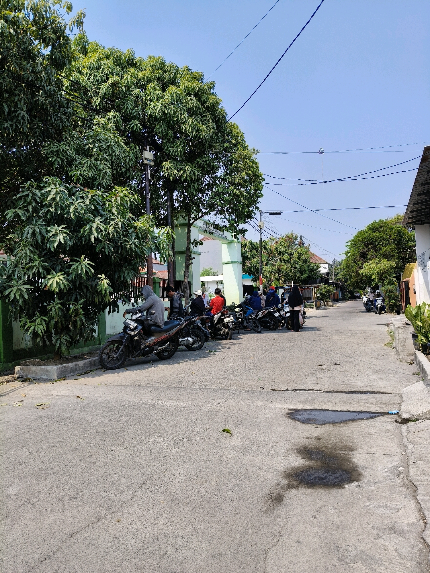 A group of motorcycles are parked on the side of a paved road in a residential area in Bekasi, Indonesia. Several people are standing or sitting on the motorcycles. A large tree with green leaves is on the left side of the image, and a building with green accents is behind the motorcycles. The sky is clear and blue. The road shows some signs of wear and tear, with small puddles of water and some oil stains visible.
