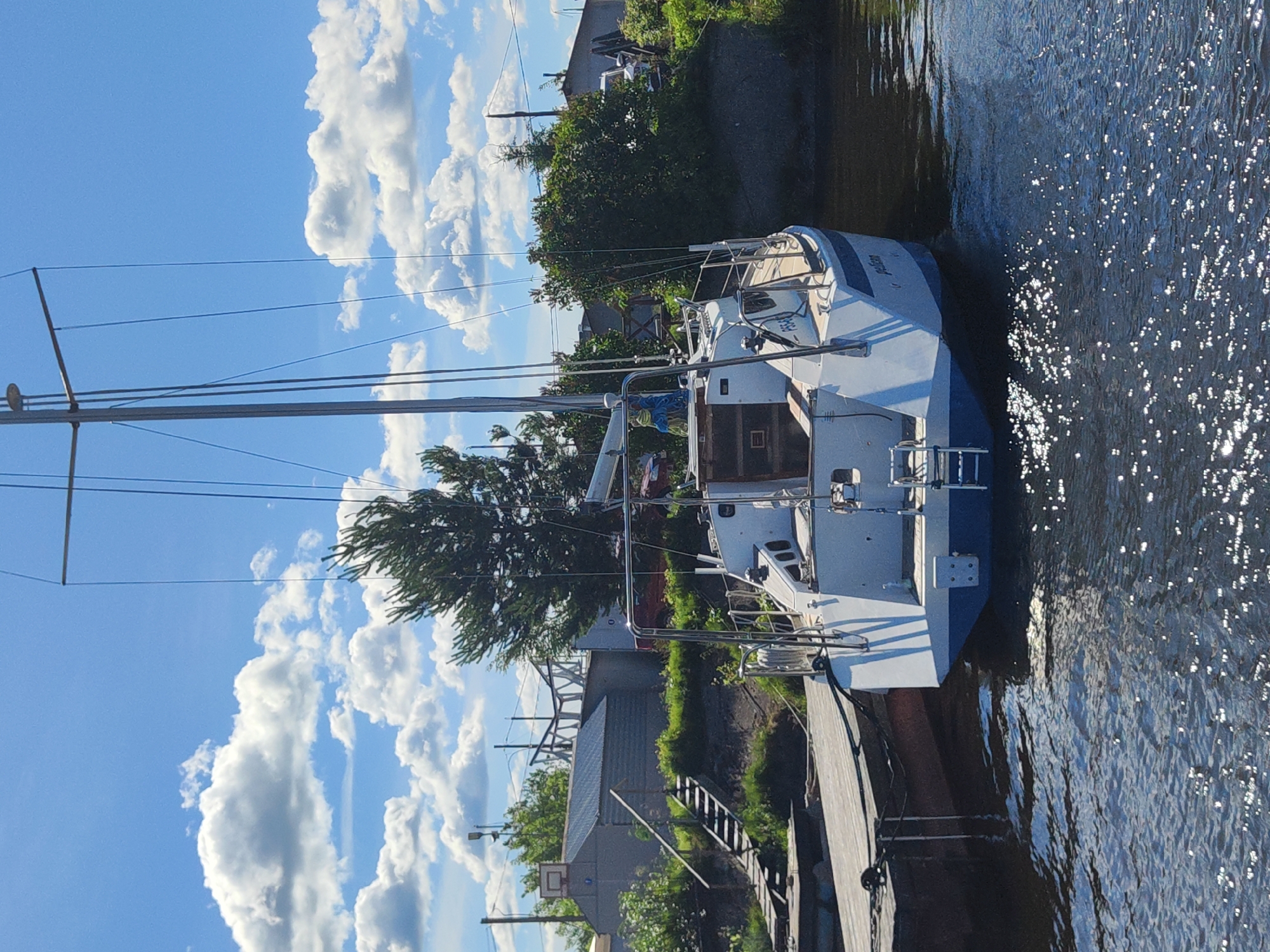 The image depicts a sailboat docked at a wooden pier on a sunny day, with fluffy white clouds scattered across a vibrant blue sky. The sailboat is white with blue accents and has a tall mast. The water is calm and reflects the sunlight, creating a shimmering effect.
In the background, there are trees and buildings, suggesting a waterfront community. One building appears to be a boathouse or storage shed, with a basketball hoop mounted on its side. The overall scene evokes a sense of tranquility and leisure, typical of a summer day by the water. There is no visible text in the image. The orientation of the image is rotated 90 degrees counterclockwise.