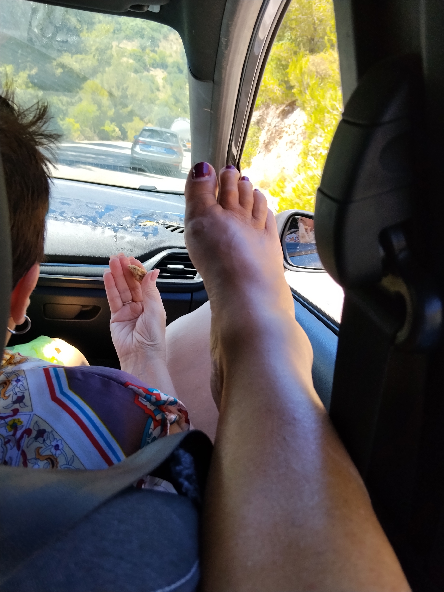 The image shows a person's bare foot, with toenails painted dark purple, resting on the dashboard of a car. The foot is positioned in the foreground, with the leg extending upwards. Through the car window, a hilly, green landscape is visible, with trees and a road where two cars are driving. The interior of the car, including the dashboard, seats, and a side mirror, is also visible. A person's hand, wearing a ring, is holding a small object, possibly food. The scene suggests a journey through a rural area in Mattinata, Italy, on a sunny day.