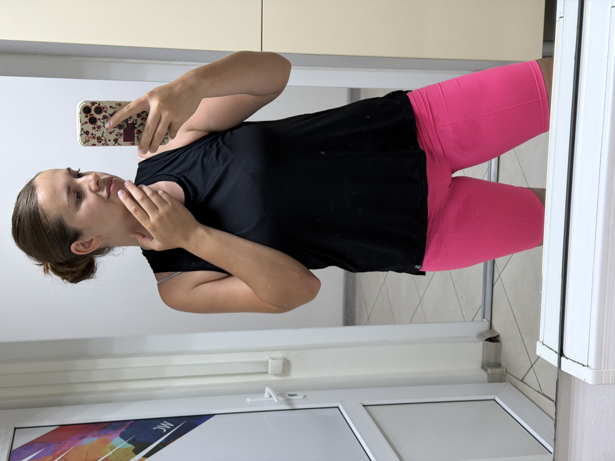 A young woman is taking a selfie in a mirror, possibly in a gym locker room or changing area. She is wearing a black tank top and bright pink biker shorts. Her hair is pulled back in a bun. She is holding a smartphone with a floral case in her right hand. The background shows a door with a colorful graphic on it, a white wall, and part of a locker. The floor is tiled. The scene is brightly lit. There is no specific indicator of the location being in Sochi, Russia.