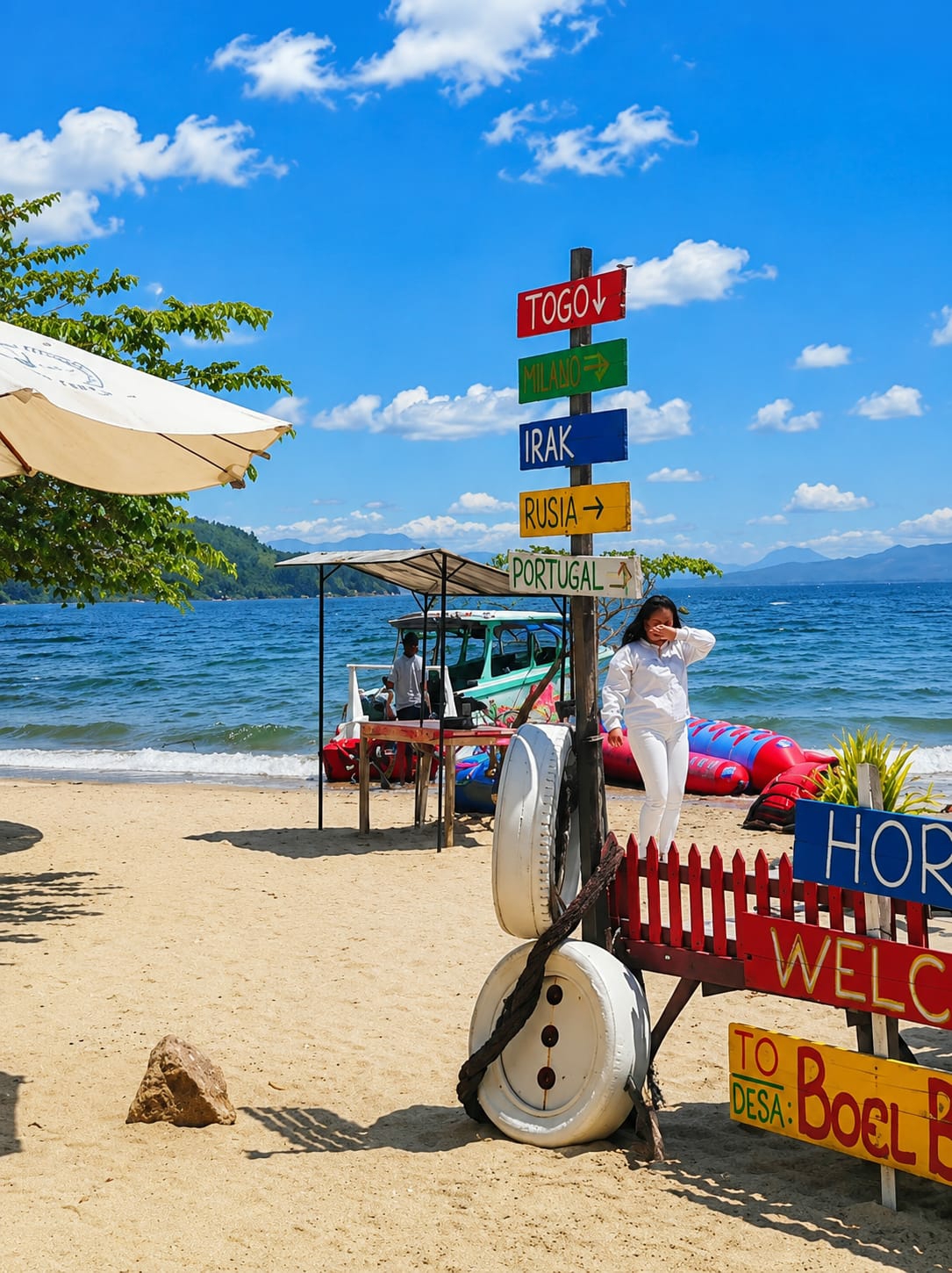 In Lumban Bul Bul, Indonesia, this photograph captures a beach scene under a bright, cloudy sky. A directional signpost stands prominently, displaying destinations such as Togo, Milano, Irak, Rusia, and Portugal. To the right of the signpost, a person dressed in white stands near a vibrant red and blue inflatable water toy. In the background, the blue ocean meets the sandy shore, with a boat and a covered stall situated on the beach. The foreground features sand, a rock, and a decorative arrangement of tires and rope.