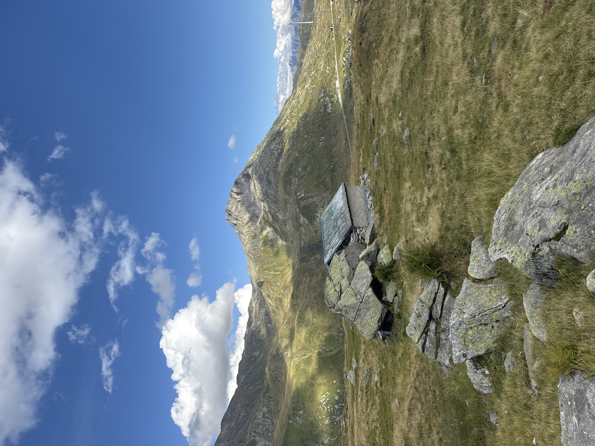 The image shows a remote mountain landscape in the Swiss Alps near Airolo, Switzerland. In the foreground, there are several large, lichen-covered boulders scattered across a grassy slope. A small, rustic stone hut with a green metal roof is situated behind the boulders, partially nestled into the rocky terrain.
The midground features rolling, green hills and mountainsides, with dramatic shadows cast by the sunlight, indicating a sunny day with scattered clouds. A road or track winds through the landscape in the distance.
The background is dominated by a large, rugged mountain peak bathed in sunlight, with a clear blue sky above dotted with white, puffy clouds. To the right, in the far distance, a modern wind turbine stands tall against the mountainous backdrop, suggesting the presence of human infrastructure in this otherwise natural setting. The environment is characterized by its alpine terrain, with abundant grass, rocks, and dramatic mountain formations. There are no people or animals visible in the frame.