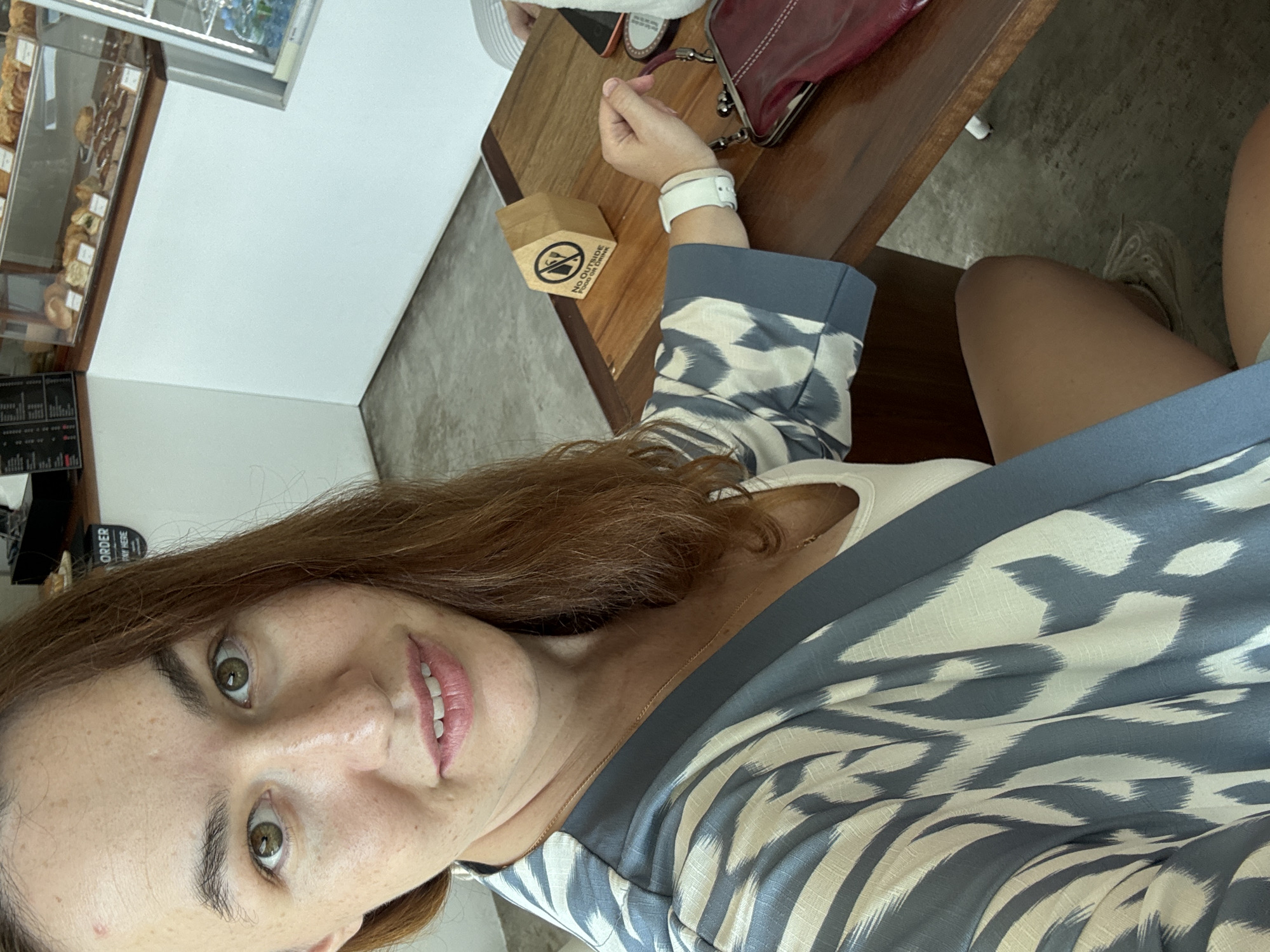 A young woman with long, auburn hair is seated at a wooden table in what appears to be a cafe in Phang-nga, Thailand. She is taking a selfie, with her face filling the foreground of the image. She is wearing a white tank top and a blue and white patterned kimono-style robe. A thin gold necklace is visible around her neck. Her right hand, adorned with a white wristband, rests on the table, holding a small maroon purse. A small, brown, house-shaped sign on the table reads "NO OUTSIDE FOOD OR DRINK." In the background, a display case filled with pastries and a counter with a menu are visible, suggesting the cafe setting. The floor is concrete.