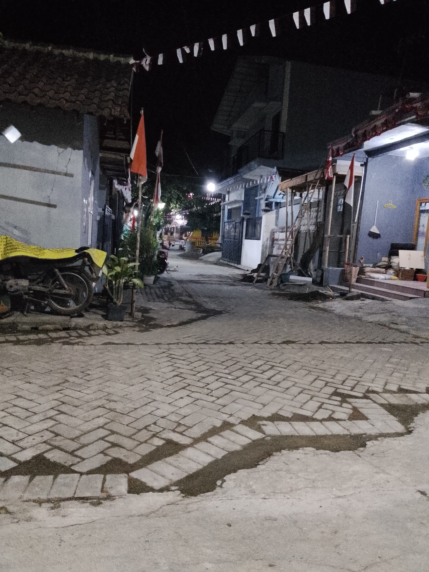 The image depicts a street scene at night in Pasuruan, Indonesia. The foreground is paved with interlocking gray bricks, with dark, irregular patches of what appears to be wet asphalt or mud. To the left, a motorcycle covered with a yellow tarp is parked next to a building with a gray facade and a tiled roof. A small potted plant stands beside the motorcycle.
The street extends into the background, lined with buildings on both sides. Several Indonesian flags, red and white, are visible, some hanging from poles and others on wires strung across the street, adorned with white triangular pennants. On the right side of the street, a building has a blue facade with a covered porch area and a set of stairs leading up. A broom and a yellow hanger are visible on the wall. Further down the street, more buildings and trees are dimly lit. The overall impression is a residential or mixed-use street decorated for a celebration or holiday.