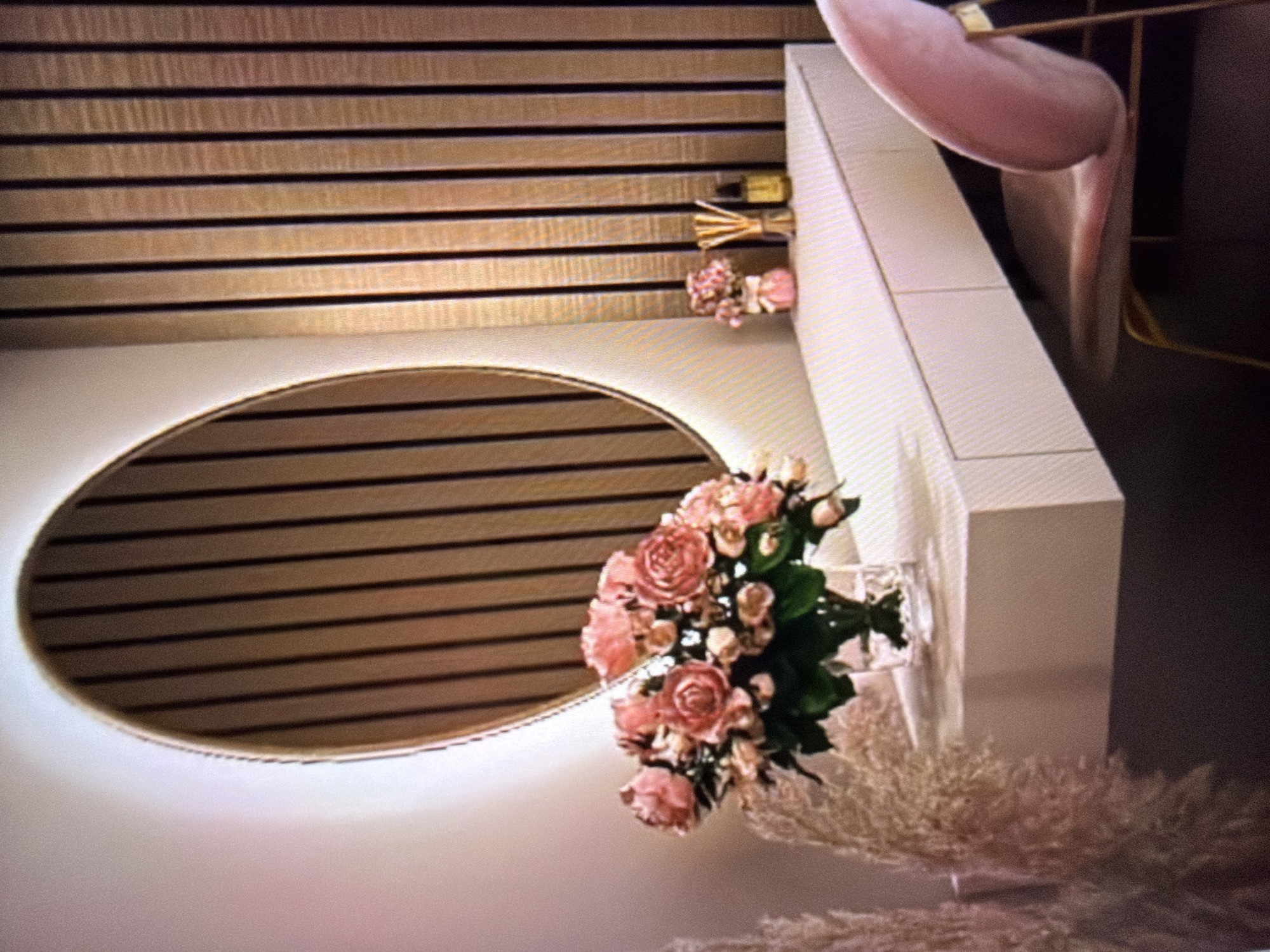 This image depicts a vanity area, likely in a residential setting, featuring a minimalist white floating vanity with subtle drawer lines. On the vanity, a clear glass vase holds a large bouquet of pink roses and smaller pink blossoms, accompanied by a small pink decorative item, a reed diffuser with golden liquid, and a small clear glass vase with reeds. Behind the vanity, a large, illuminated oval mirror with a fluted wood backing is mounted on a wall adorned with vertical wooden slats. To the left of the vanity, tall, wispy, pale pink decorative reeds are visible. A plush pink velvet chair with gold-toned legs is positioned to the right of the vanity. The overall scene suggests a modern and elegant dressing or beauty station. The image does not provide any visual cues or information to ascertain its location in Beșalma, Moldova.