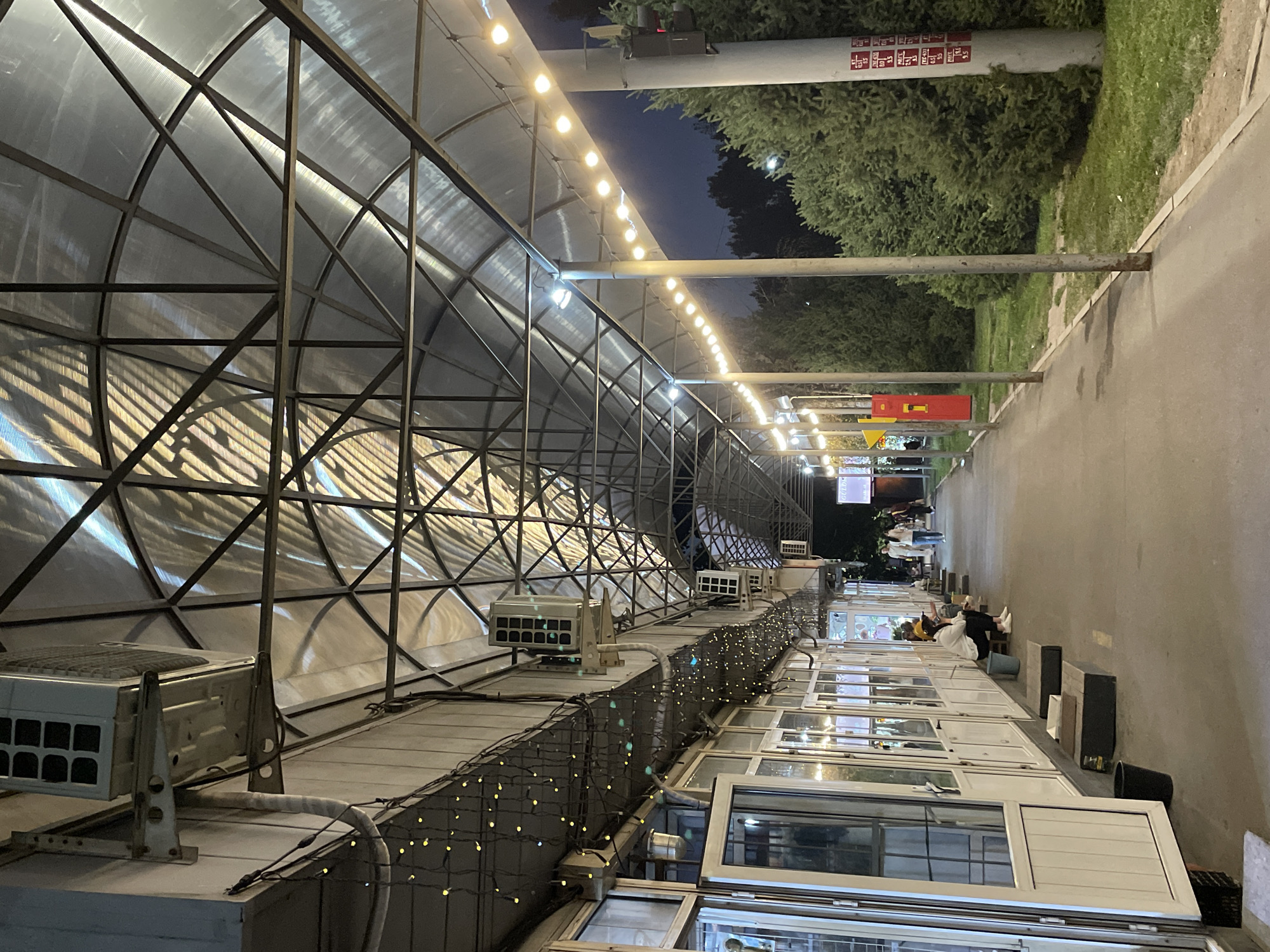 The image depicts an outdoor seating area under a transparent, arched canopy supported by metal beams. The canopy is illuminated by strings of small yellow lights and several larger, bare bulbs. Along the left side of the walkway, there are several shops or restaurants with large glass windows and white doors, some of which are open. Air conditioning units are mounted on the walls above the shops. A few people are seated on wooden benches or on the ground outside the establishments. The walkway extends into the distance, lined with trees and tall poles with more lights. On the right side of the walkway, there is a grassy area with dense evergreen trees and a lamppost with some signage attached. The overall scene appears to be a public space, possibly a pedestrian alley or a market area, at dusk or night. The presence of air conditioning units and the style of the buildings suggest an urban environment. This scene is consistent with the outdoor dining and market areas found in Almaty, Kazakhstan, particularly along streets with commercial establishments and pedestrian walkways.