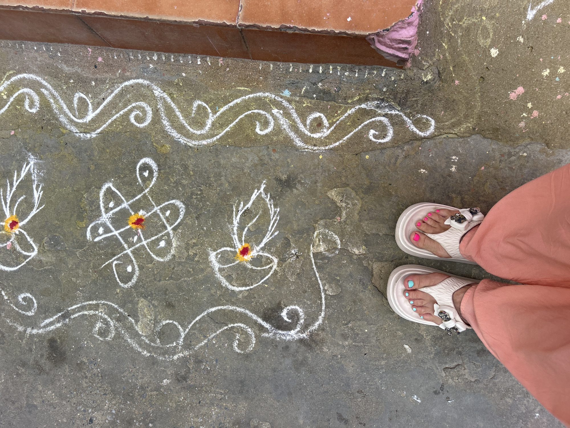 The image shows a chalk-drawn Kolam design on a concrete surface, likely near a building or entrance, based on the tile edging at the top of the frame. The Kolam features intricate swirls, geometric patterns, and lamp-like figures, with the design highlighted by added yellow and red coloring at the center of the geometric figures and above the lamp figures. A pair of feet in white sandals with floral embellishments and brightly painted toenails (pink and blue) are visible in the frame, with the person wearing coral-colored pants. The setting is most likely Puttaparthi, India, indicated by the Kolam art, which is a traditional South Indian art form often seen in front of homes and businesses. The surface has some cracks and discoloration, suggesting it's an outdoor, publicly accessible area.