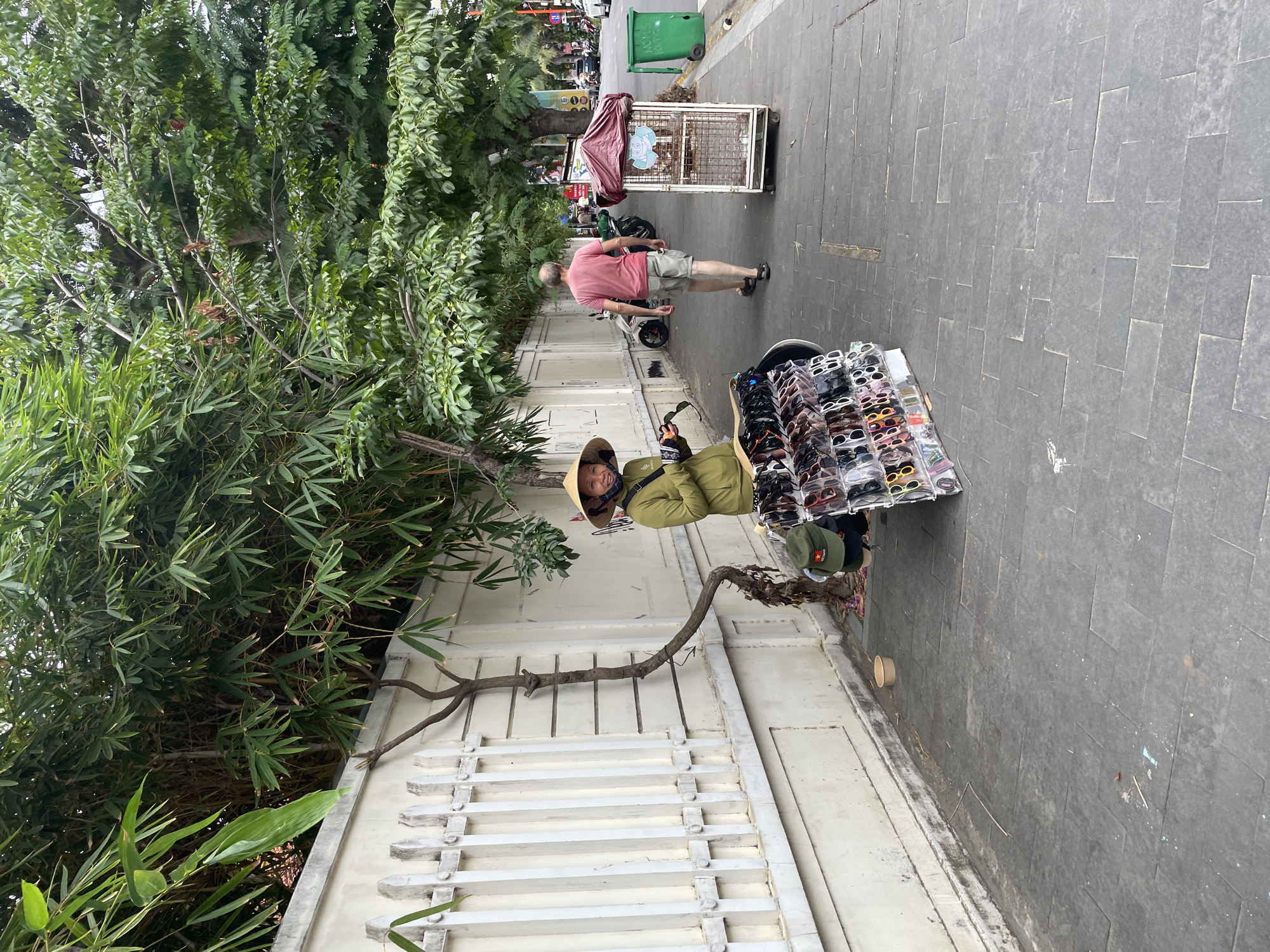 A street vendor wearing a conical hat and a green jacket stands beside a display of sunglasses and hats on a sidewalk in Da Nang, Vietnam. Behind the vendor is a white wall with vertical slats, bordered by bamboo plants and trees. Further down the sidewalk, a man in a pink shirt and shorts walks away from the camera. A caged cart with a red canopy sits on the sidewalk, and a green trash bin is visible in the background. The street is paved with dark gray tiles, and there are trees lining the side of the road, casting shadows on the sidewalk. Cars and motorcycles are present on the street, indicating an urban environment.