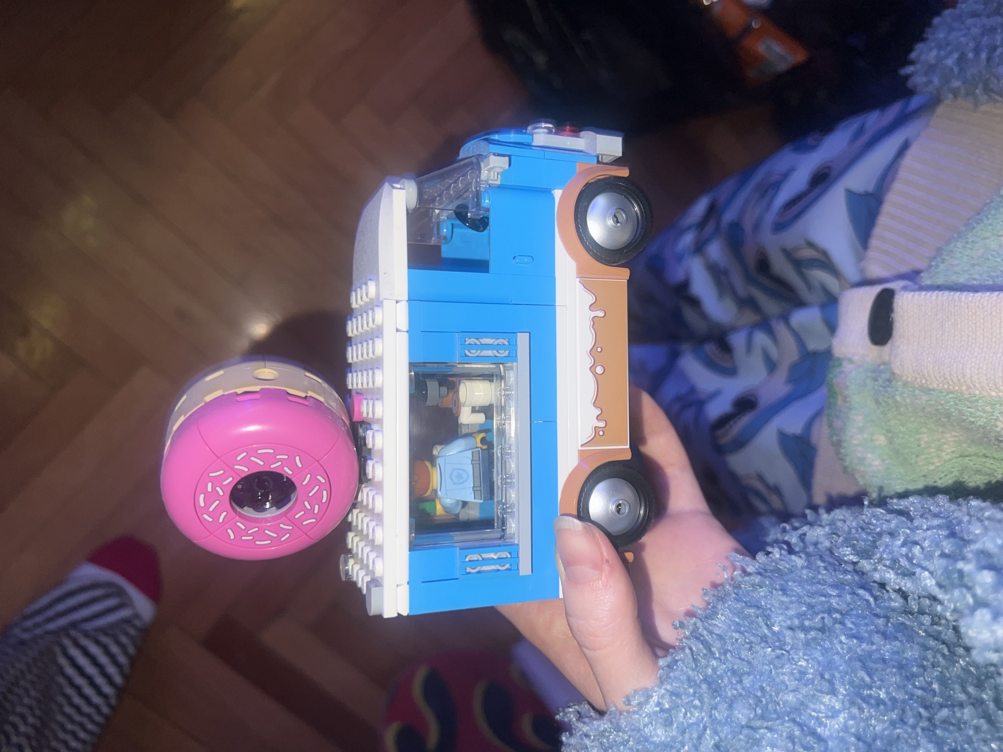 A person holds a Lego donut shop truck in their hand. The truck is blue and white with brown accents to mimic frosting, and a pink donut sign is attached to the side. The truck's window shows Lego minifigures inside. The background includes a hardwood floor, patterned pajamas, and a blue fuzzy blanket. The location is not possible to identify based on visual data, so I cannot specify that it is in Belgrade, Serbia.