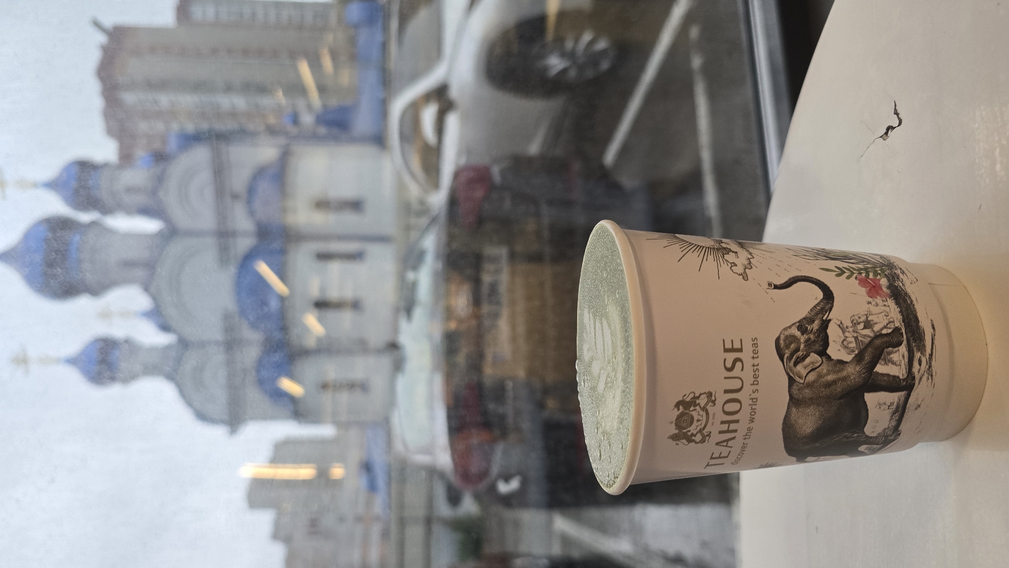 In the foreground, a paper cup with the "TEAHOUSE" logo and an illustration of an elephant is placed on a white surface. The cup contains a green beverage with frothy latte art and water droplets on its surface. In the background, viewed through a window, a blurred cityscape is visible. The cityscape includes a building with blue domes resembling a church, modern buildings, and parked cars. The weather appears to be rainy, as indicated by the water droplets on the window and the wet appearance of the cars.