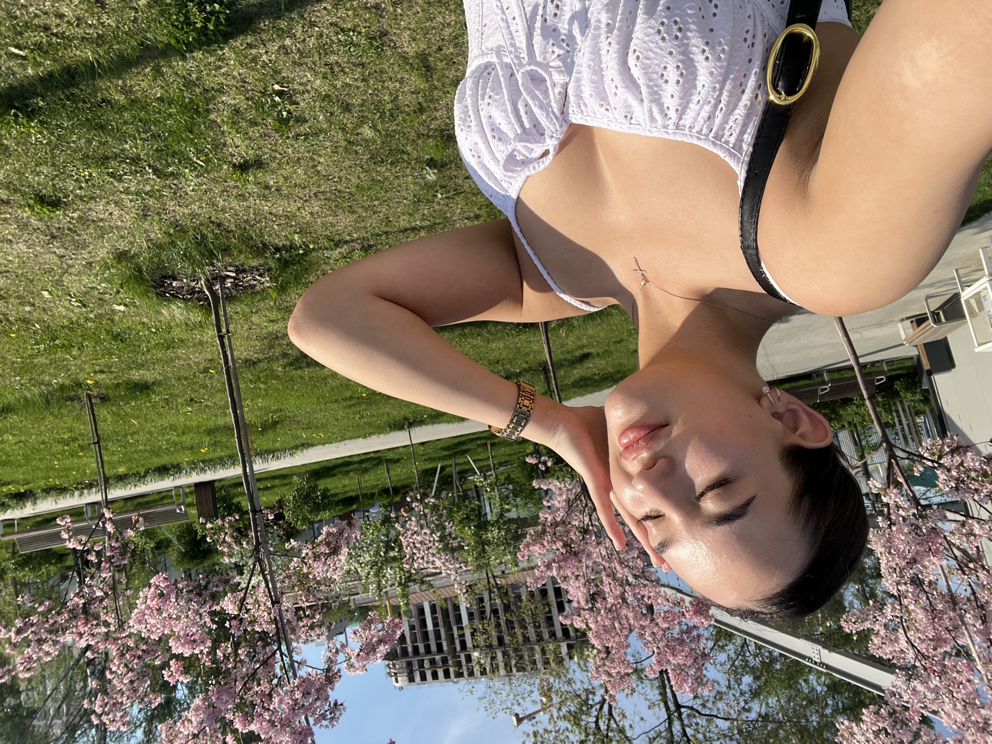 A woman poses for a selfie outdoors in Perm, Russia. She is lying in the grass with flowering trees around her and a building under construction in the background. The woman is wearing a white eyelet top and a black leather purse with gold hardware. Her left hand is near her face and she is wearing a gold watch on her wrist. She has a cross necklace. The grass and trees suggest the location is a park. The selfie is taken in an area of the city that has blossoming trees, possibly an orchard or landscaped parkway within the urban setting.