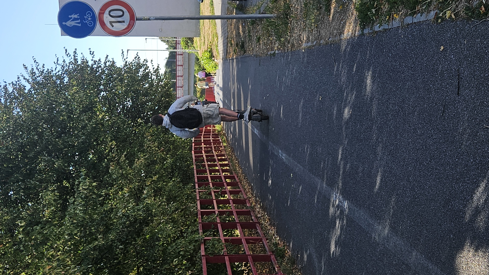 The image captures a sunny day on a paved path in Prešov, Slovakia, likely a designated bicycle and pedestrian lane. A young man, seen from behind, rides an electric scooter, wearing a gray hoodie, light-colored shorts, and a black backpack. The path is bordered on the left by a bright red metal fence and dense green foliage. On the right side of the path, a traffic sign indicating a speed limit of 10 km/h for bicycles and pedestrians is visible. Further in the background, two women walk away from the camera, and a bridge spans overhead. The ground is asphalt, with scattered leaves and shadows from trees dappling the surface.