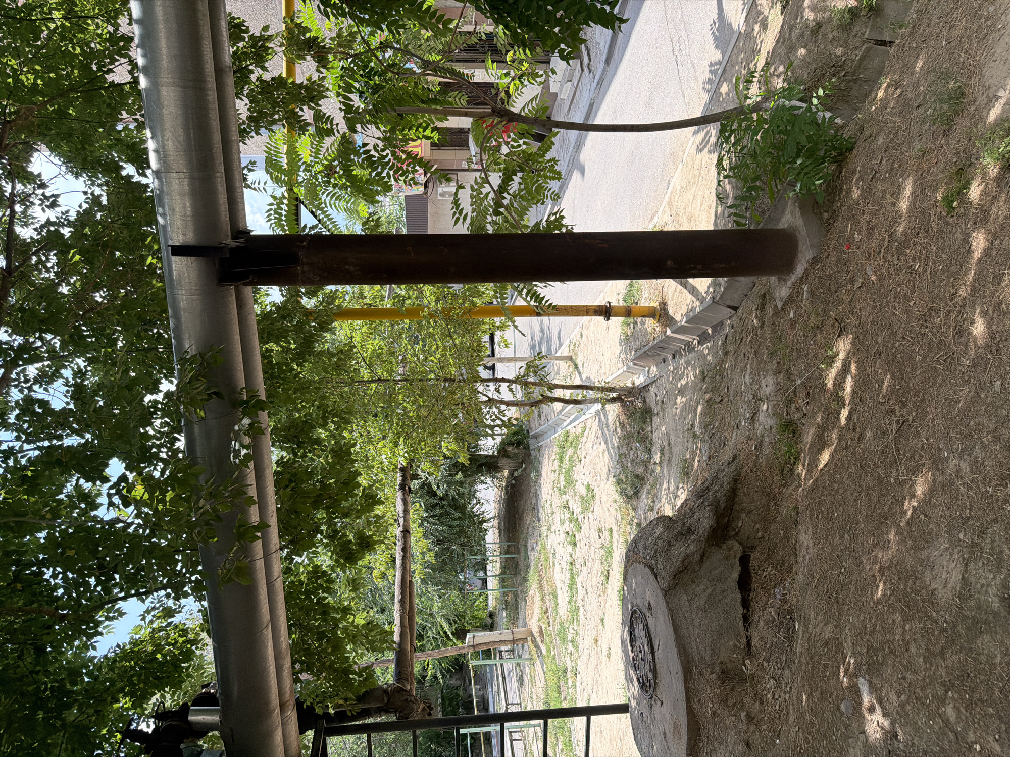 The image depicts an outdoor scene in the city of Shymkent, Kazakhstan, characterized by a dry, dusty environment with sparse vegetation and overhead utility pipes. In the foreground, a large, concrete structure resembling a tree stump with a manhole cover is visible. Beyond this, a sandy, uneven terrain leads towards a paved road with a concrete curb. Several trees with green foliage are scattered throughout the area, providing some shade. A tall, dark cylindrical pole stands prominently in the middle ground, near a yellow pole. In the background, buildings with beige and yellow facades are visible, along with a portion of the sky. The overall impression is one of an urban environment with elements of infrastructure and nature coexisting.