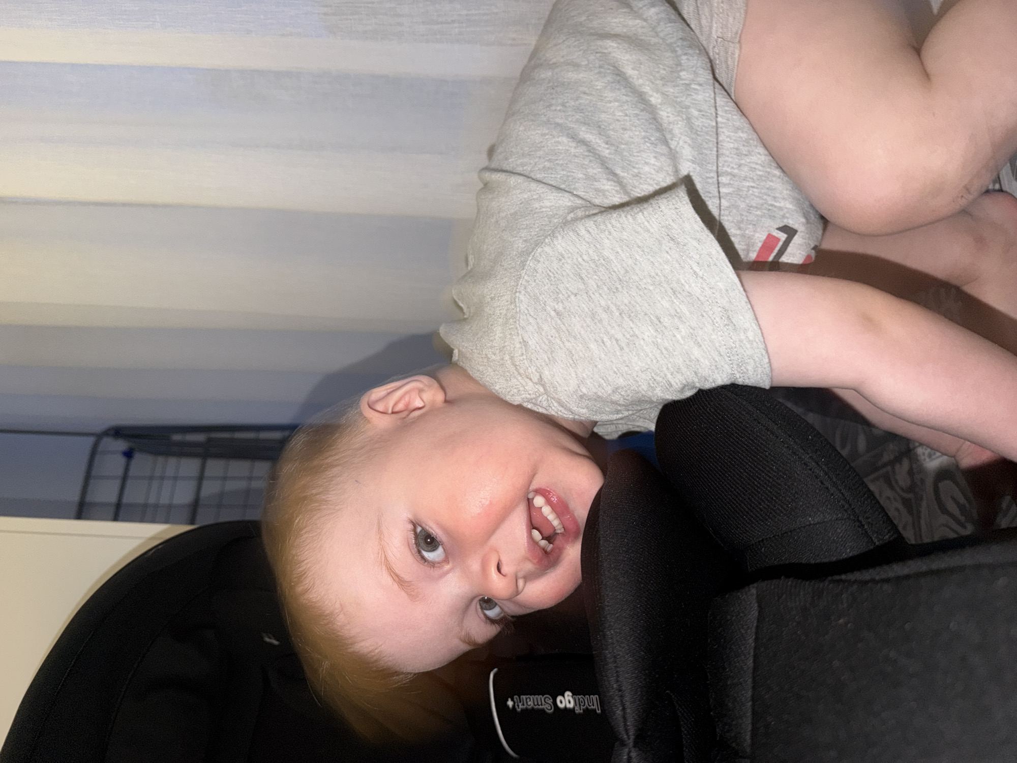 A baby is positioned upside down, angled towards the camera. The baby has fair skin, light blonde hair, and is smiling, revealing several teeth. The baby is wearing a light gray shirt. The baby is supported by a black object which looks like part of a car seat or stroller. Behind the baby are blinds and a white cabinet, and a metal rack. The location is not determinable based solely on the image. The city of Votkinsk, Russia cannot be verified from the image.