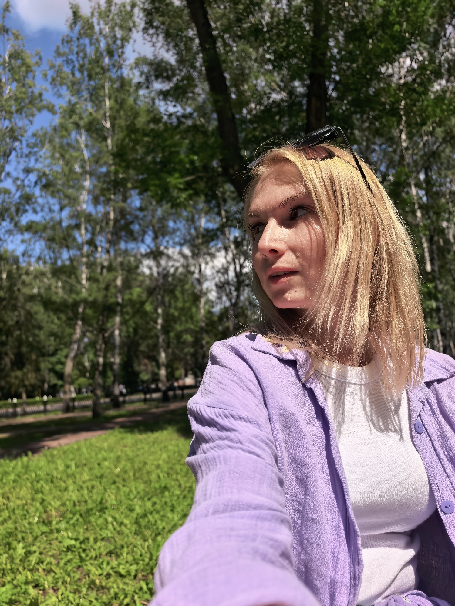 The image depicts a young woman with blonde hair, wearing a white t-shirt and an open purple shirt. She has sunglasses perched on top of her head. The woman is outdoors, with a background of green grass, trees, and a blue sky with some clouds. The overall scene appears to be a park or a natural area. There is no indication that this location is in Sterlitamak, Russia.